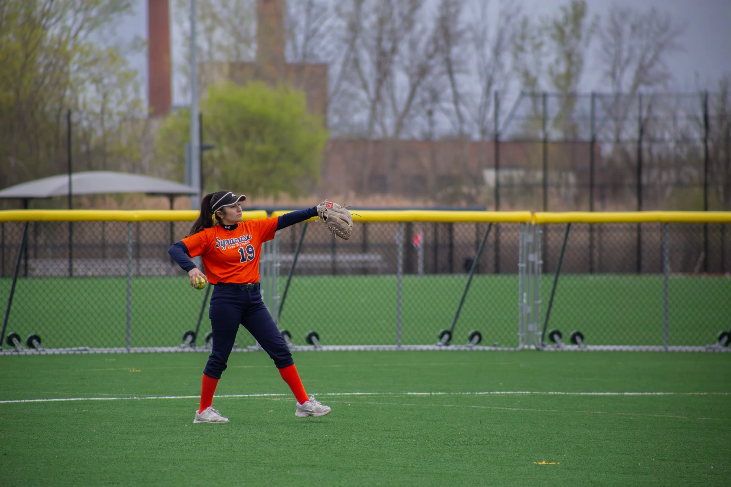 A female softball player wearing an orange jersey with the number 19, extending her arm to catch a ball on a softball field.