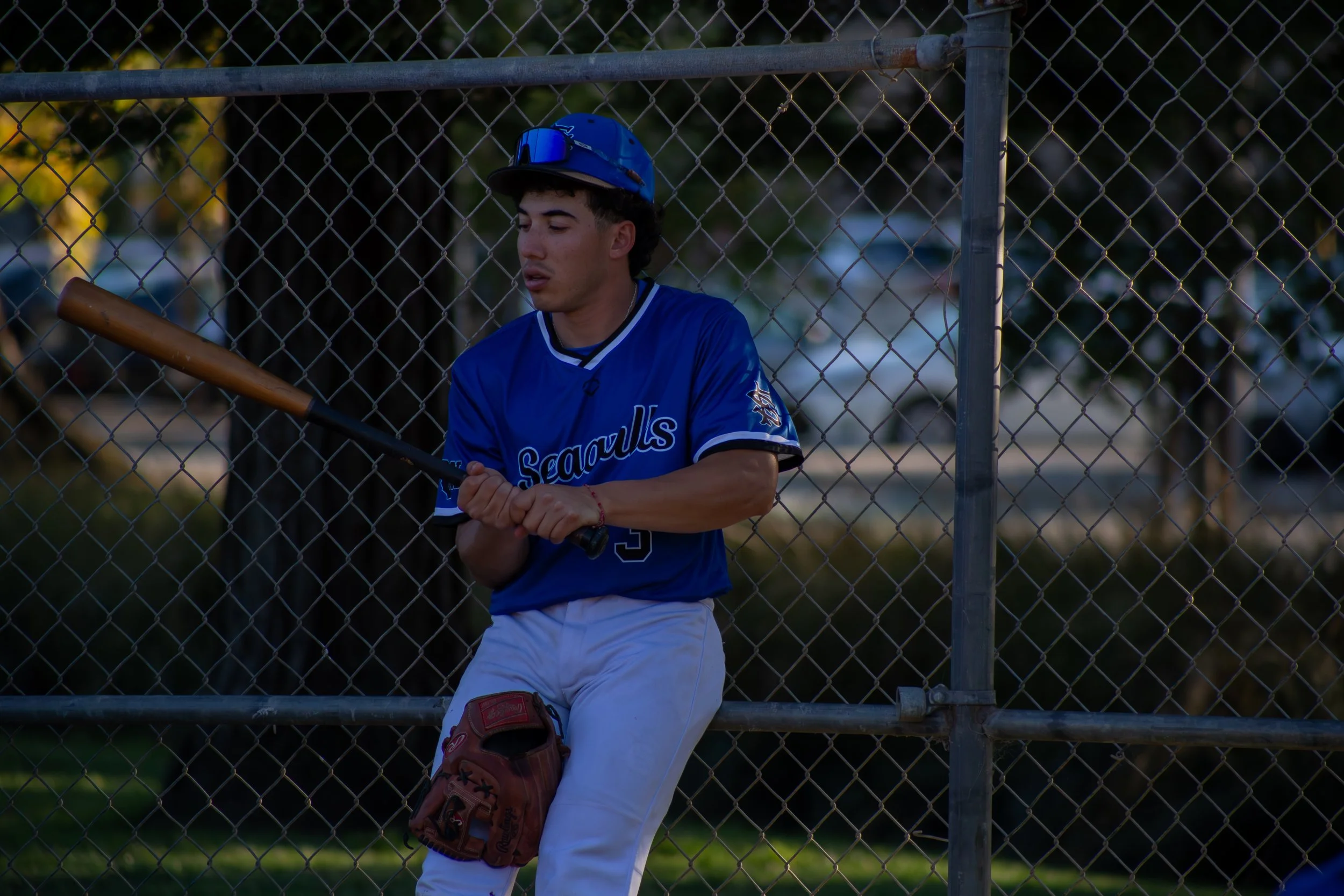 Young male baseball player sitting on a bench behind a chain-link fence, wearing a blue jersey and helmet, with a bat in his hands and a glove on his knee.