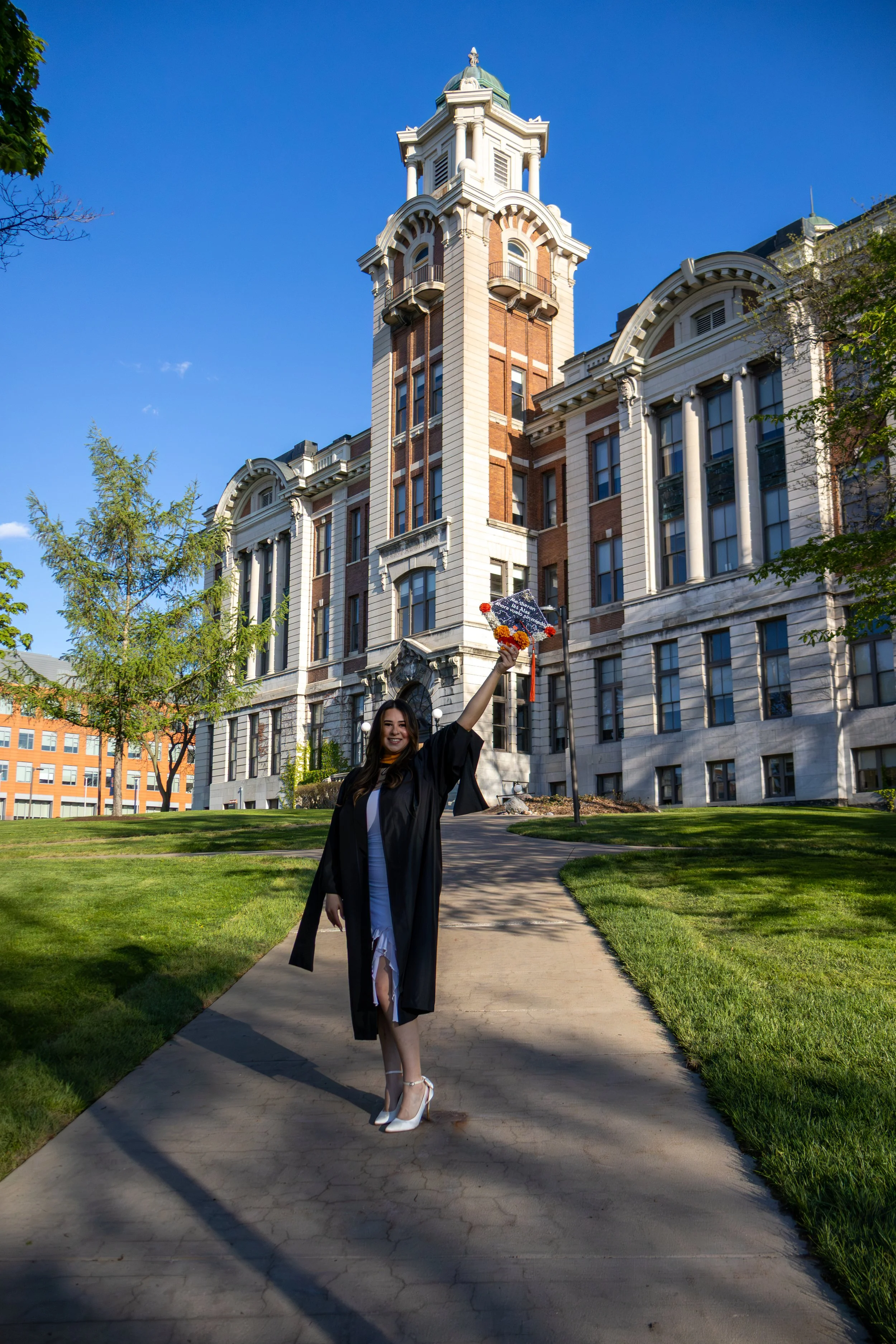 A young woman in a graduation gown and cap holding a bouquet of flowers, standing on a sidewalk in front of a historic building with a clock tower and a clear blue sky.
