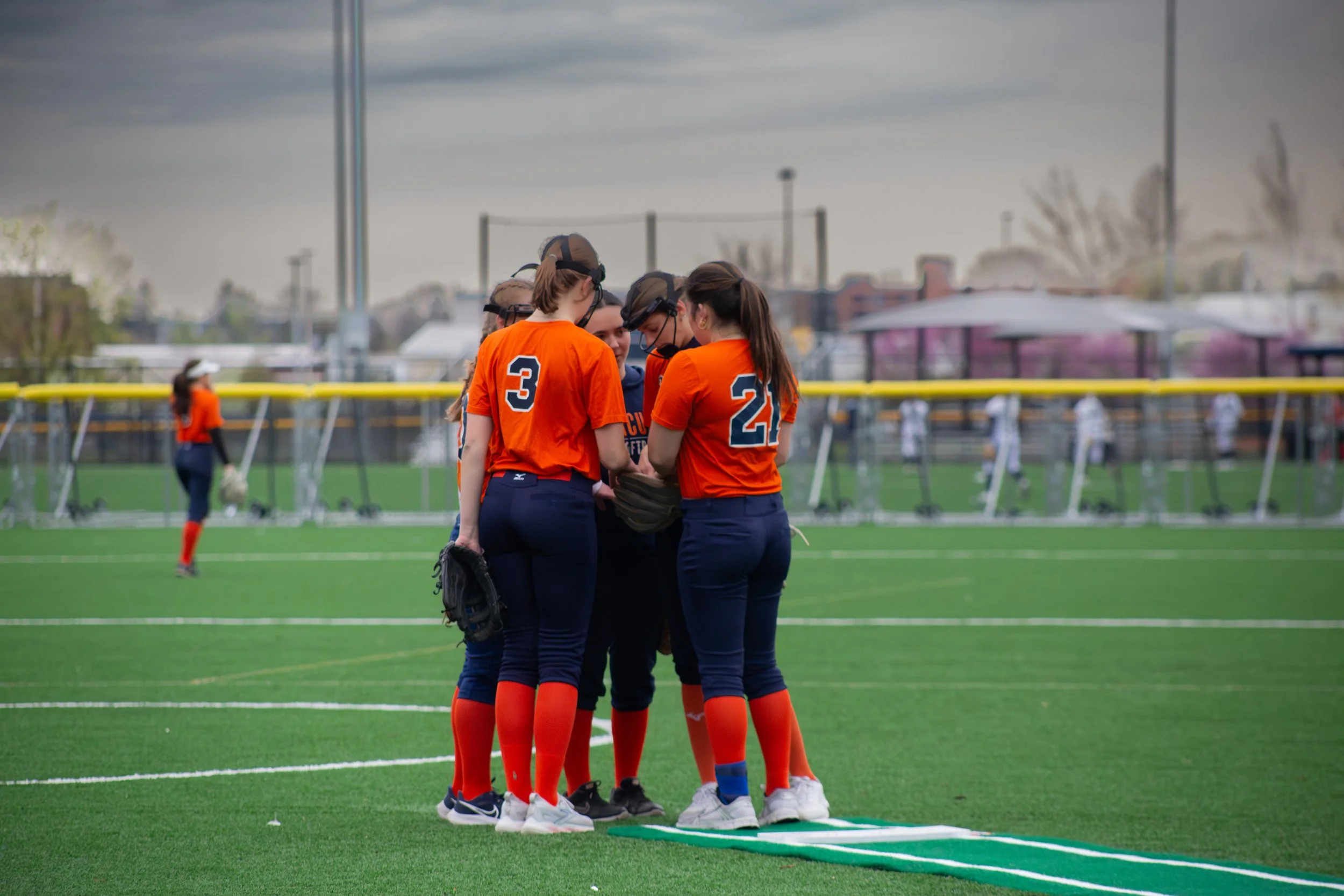 A group of softball players in orange uniforms huddle together on the field during daytime with cloudy skies.