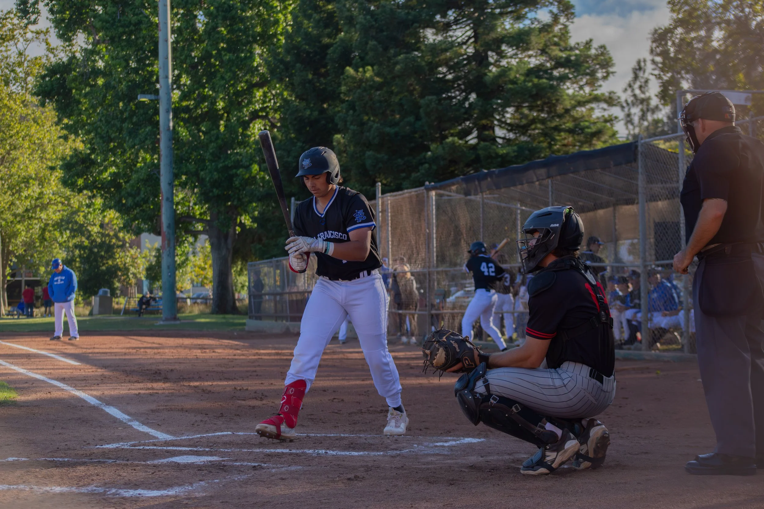 A young baseball player with a helmet and bat stands at home plate during a game. An umpire is crouched behind him, ready to catch the ball, while a coach watches on. Other players and a person in a blue jacket are visible in the background, with tre