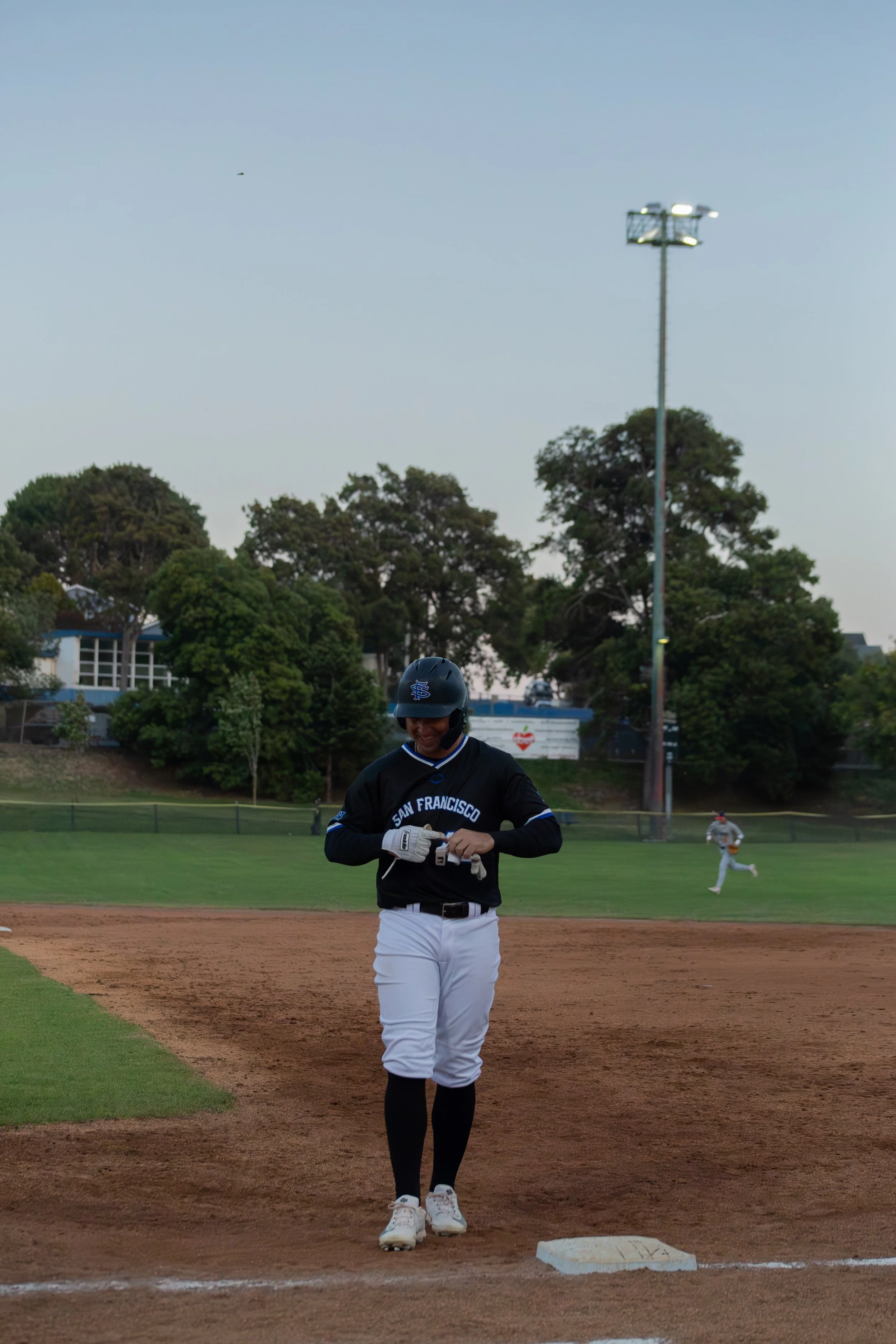 A baseball player standing at home plate on the field, wearing a black jersey with white pants, and a helmet with a 'SF' logo, smiling while holding batting gloves. In the background, another player is running on the field, with trees and a light in 