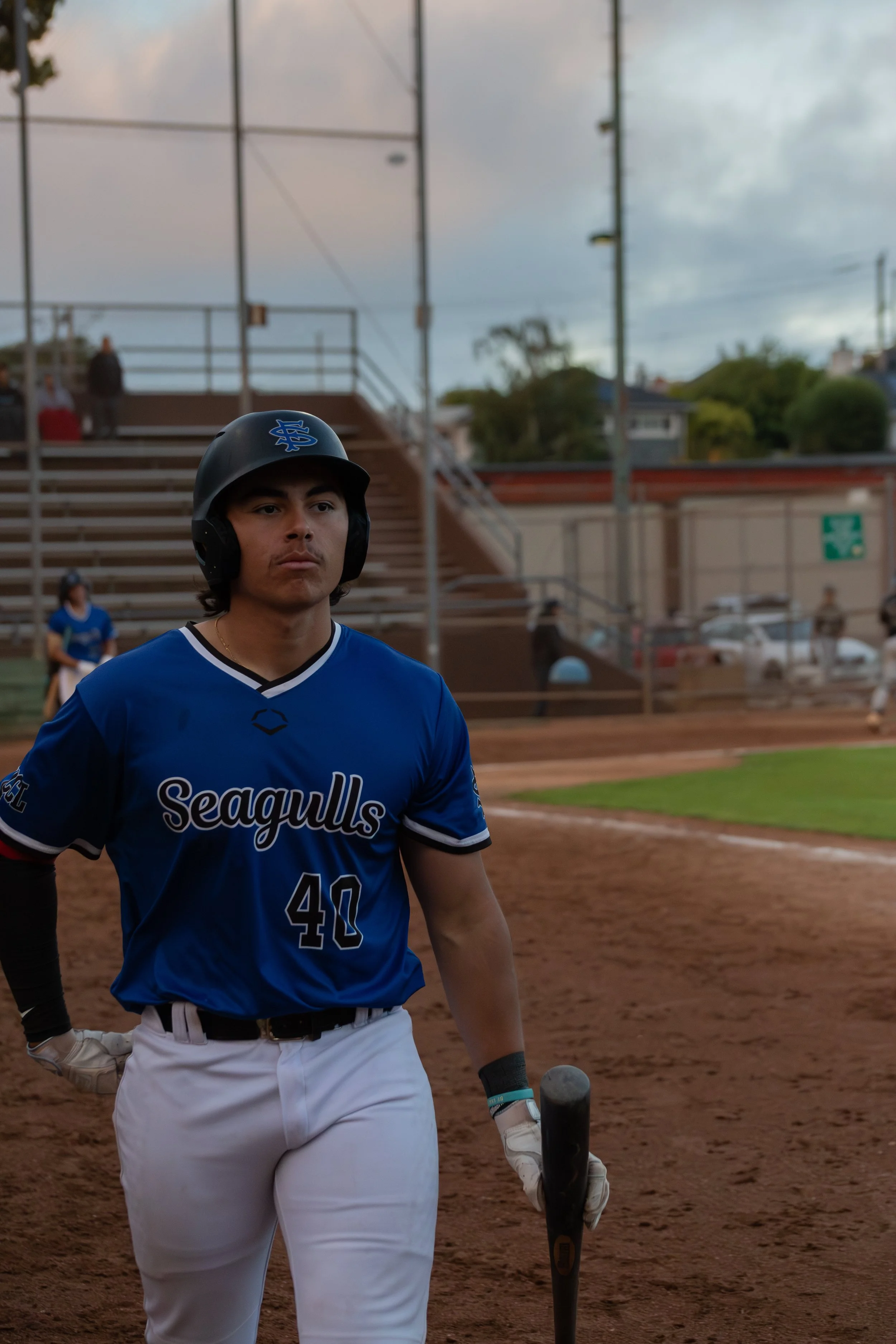 A baseball player in a blue jersey with 'Seagulls' written on it, wearing a black helmet with a logo, standing on a dirt baseball field holding a bat, with stadium bleachers and cloudy sky in the background.
