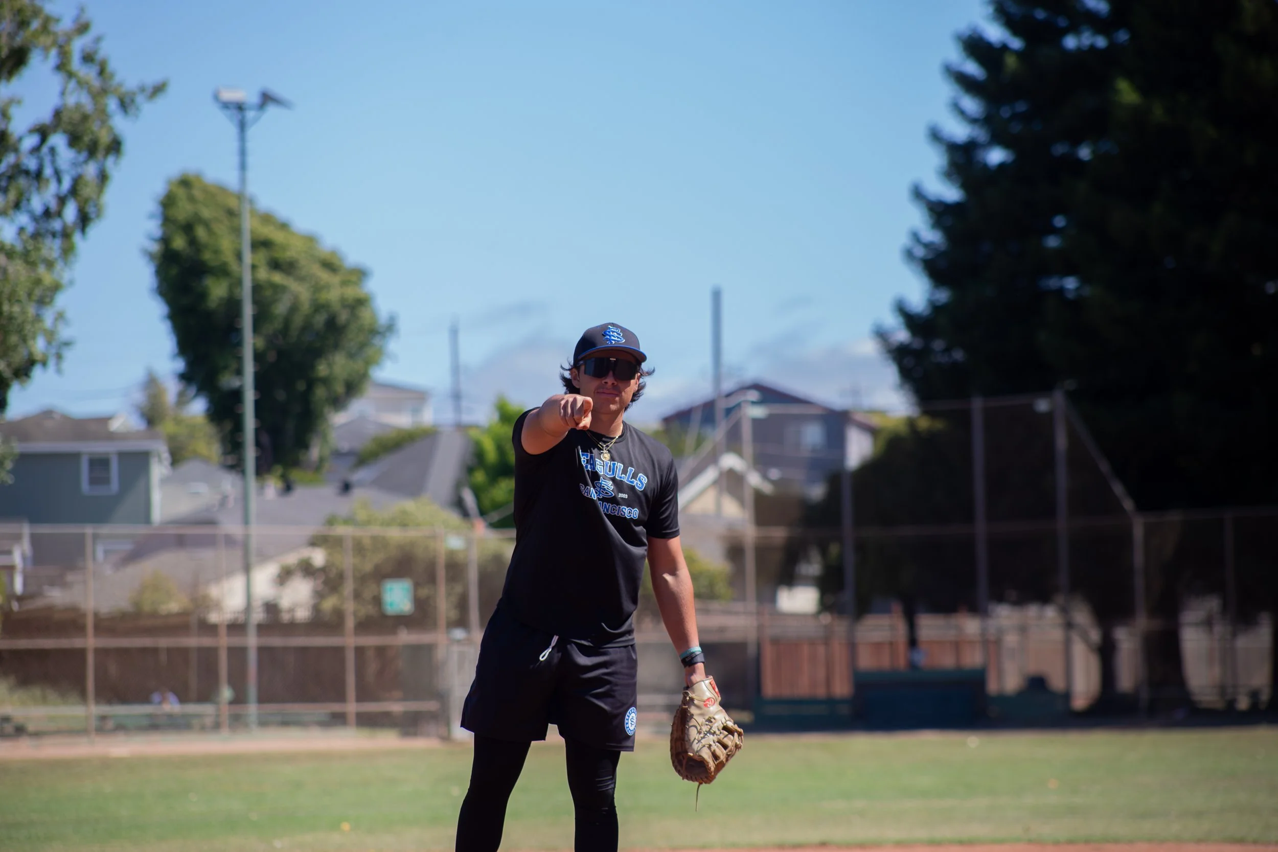 A young male baseball player pointing at the camera, wearing a black baseball uniform with blue and white lettering, standing on a grassy field with houses and trees in the background under a clear blue sky.