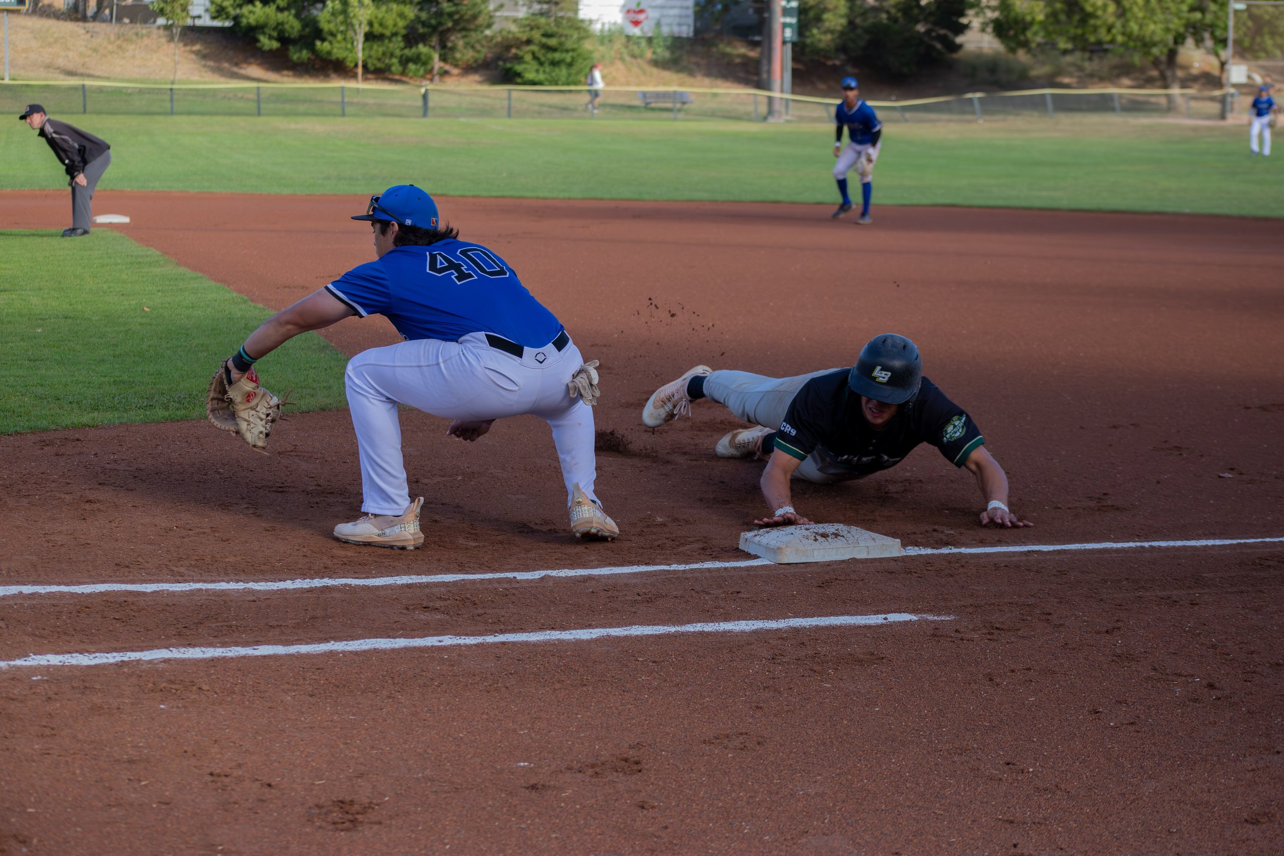 In this baseball game scene, a player wearing a blue uniform with the number 40, is diving and sliding into base while another player, also in black helmet and uniform, attempts to tag him out. An umpire watches the play closely as other players and 
