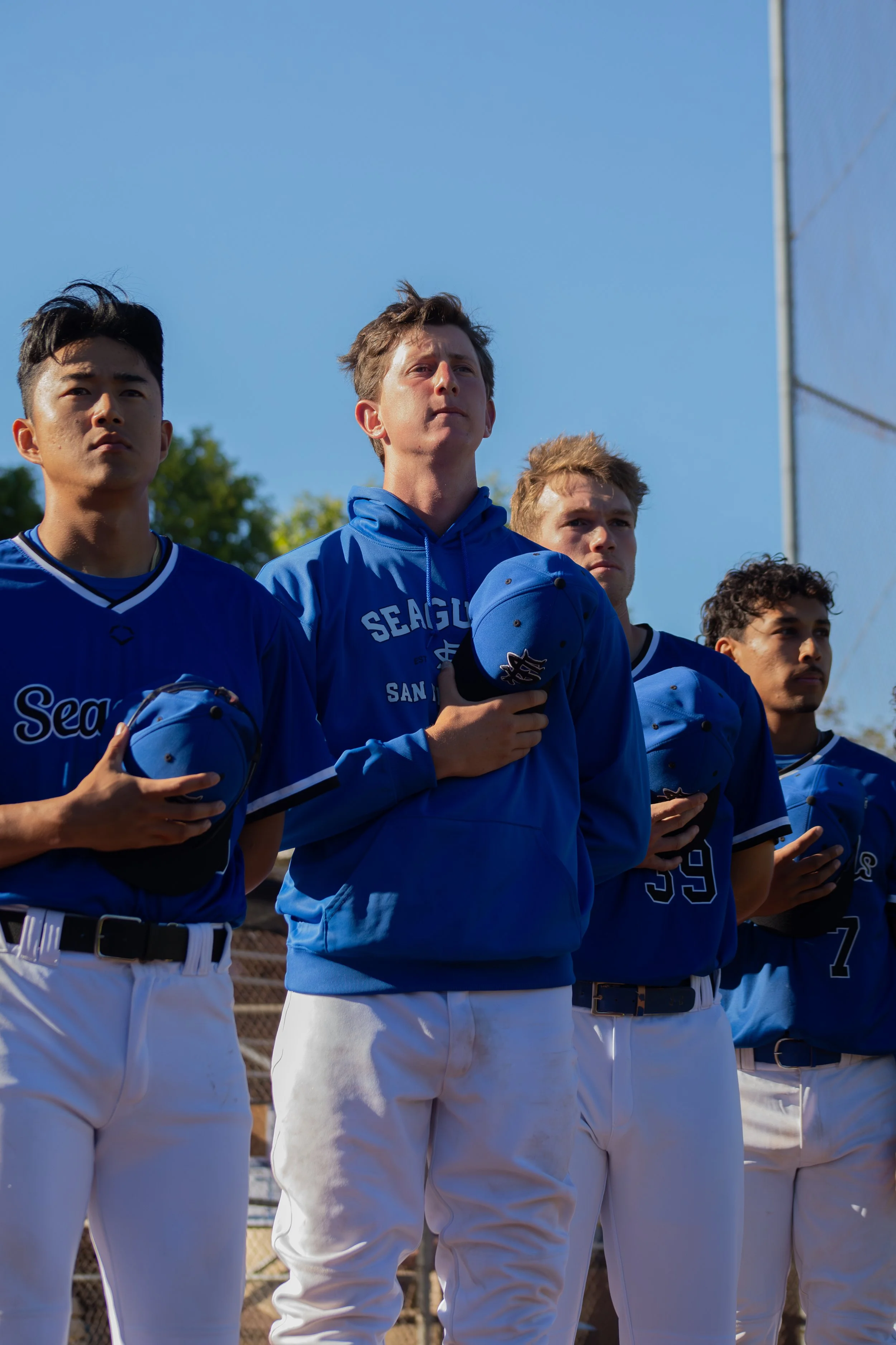 A group of baseball players standing in a line, wearing blue jerseys and white pants, with some holding baseball caps, during daylight hours.