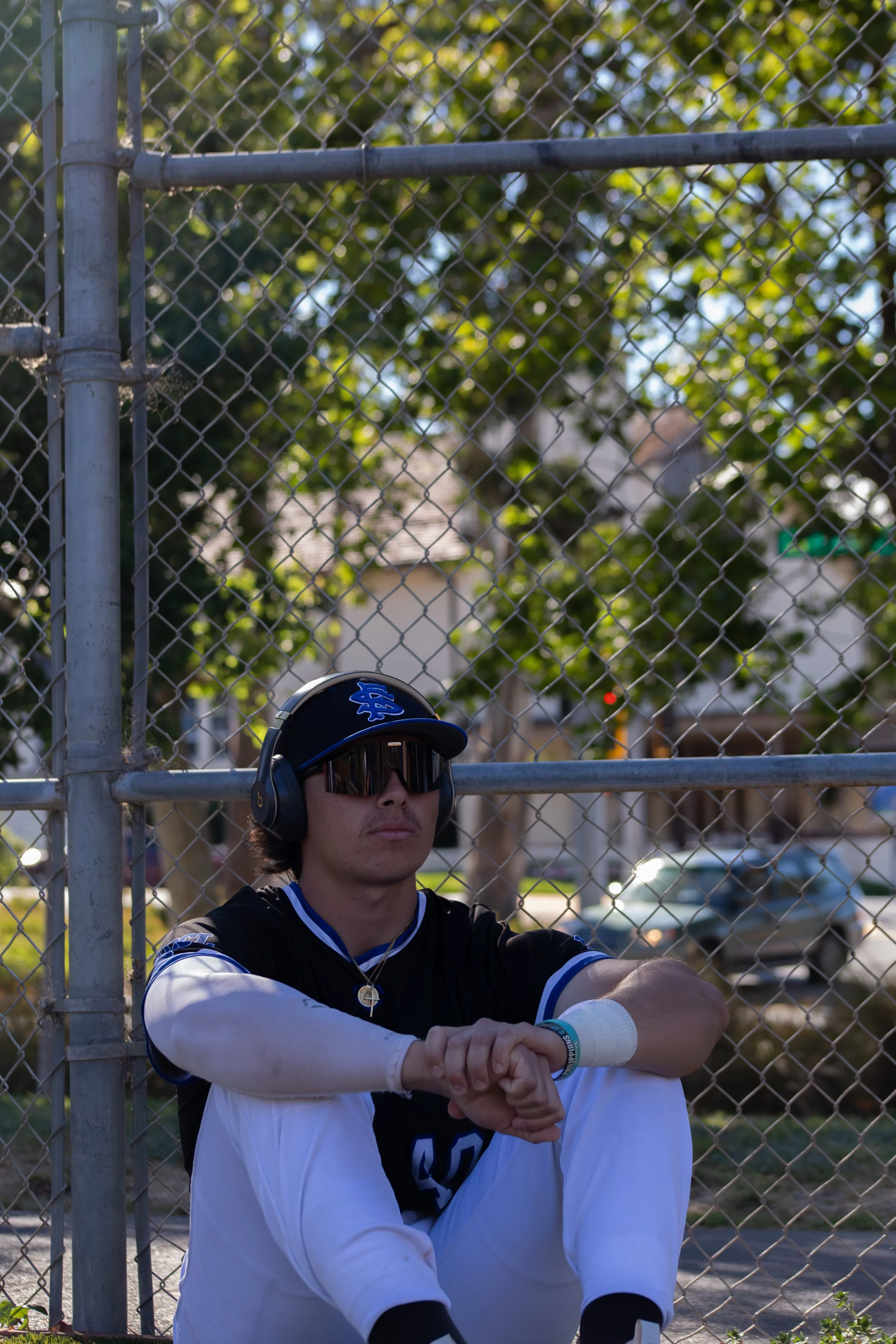 A young man with sunglasses, a baseball cap, and headphones sitting behind a chain-link fence, with trees and houses in the background.