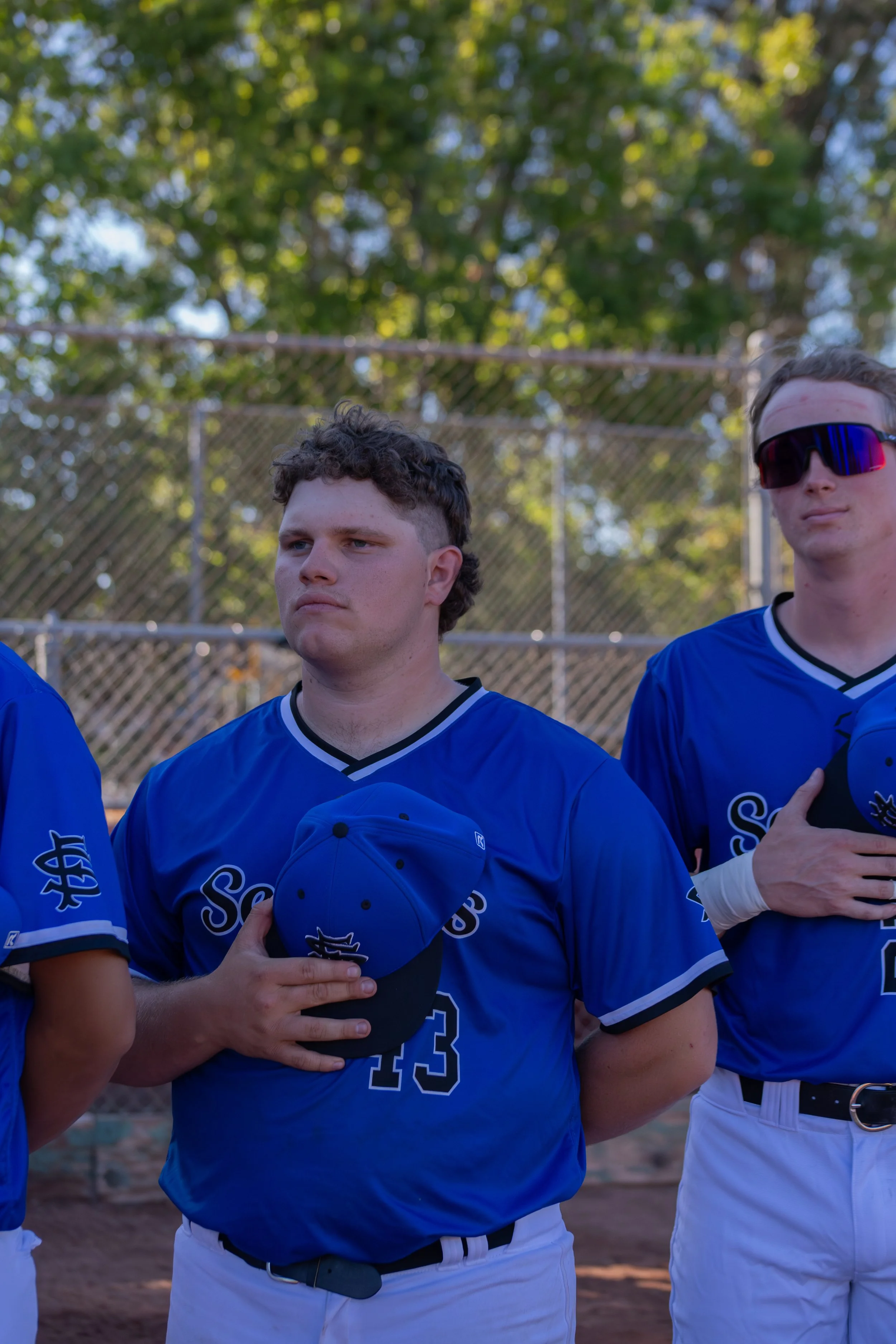 Two young men in blue baseball jerseys standing on a baseball field with a chain-link fence and trees in the background. One is holding a baseball cap, and the other is wearing sunglasses. They are standing for a photo, likely before or after a game.