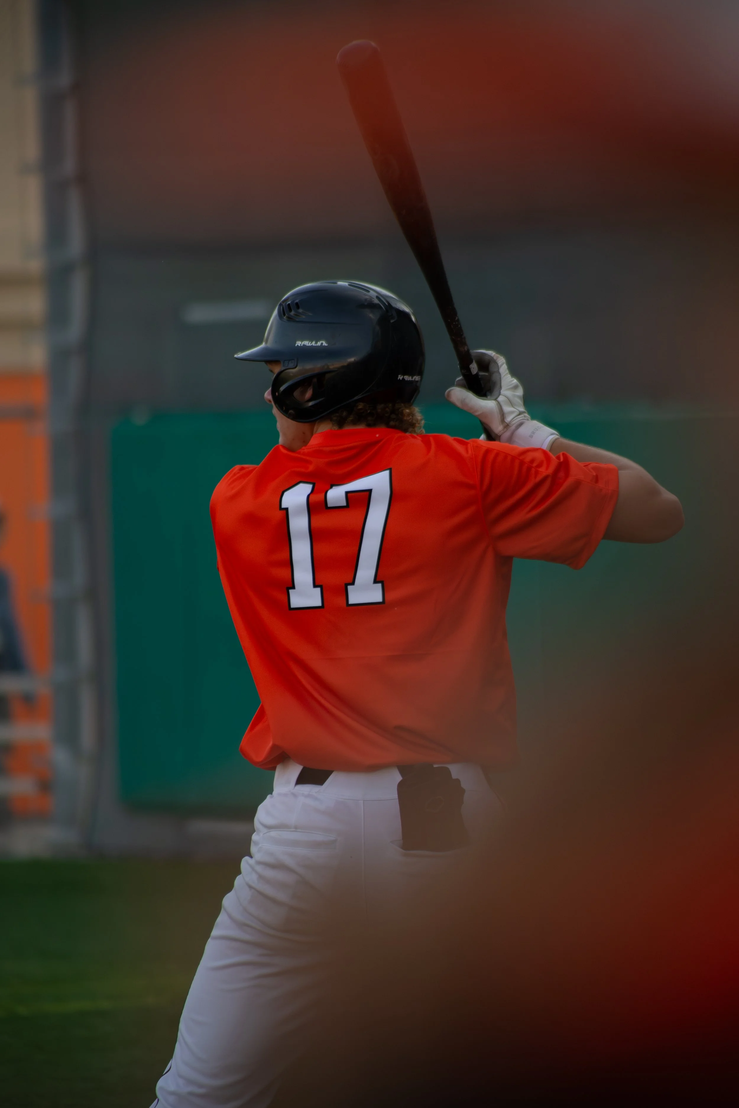 A baseball player wearing a black helmet and red jersey with the number 17 on the back is swinging a bat, preparing to hit a pitched ball. The background is blurred, focusing on the player.
