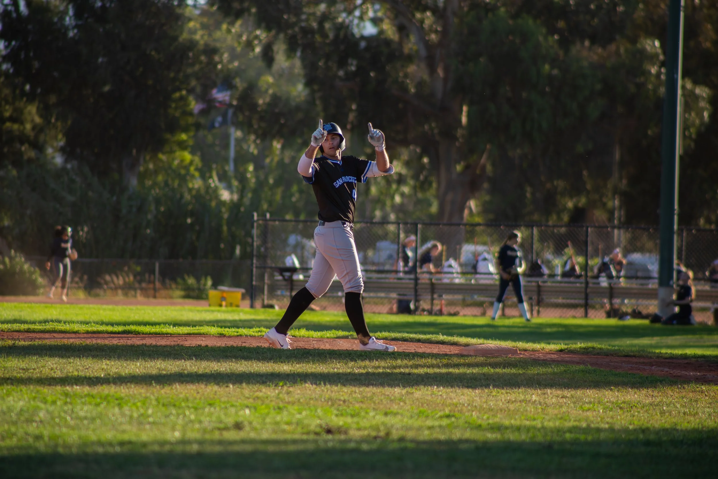 A female softball player in black and white uniform standing on the base path, looking in the distance with her finger raised, during a game at a park with trees in the background.