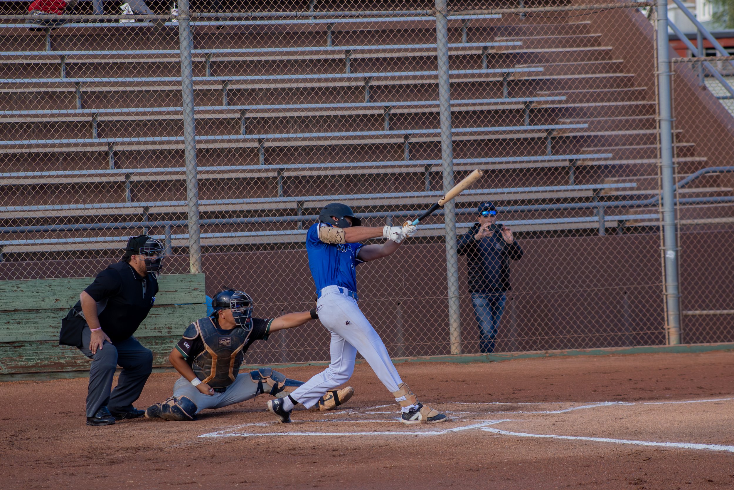 A baseball player swinging at a pitch, with a catcher and umpire crouched behind him on the field, and a woman in sunglasses taking photos behind a chain-link fence in the background.