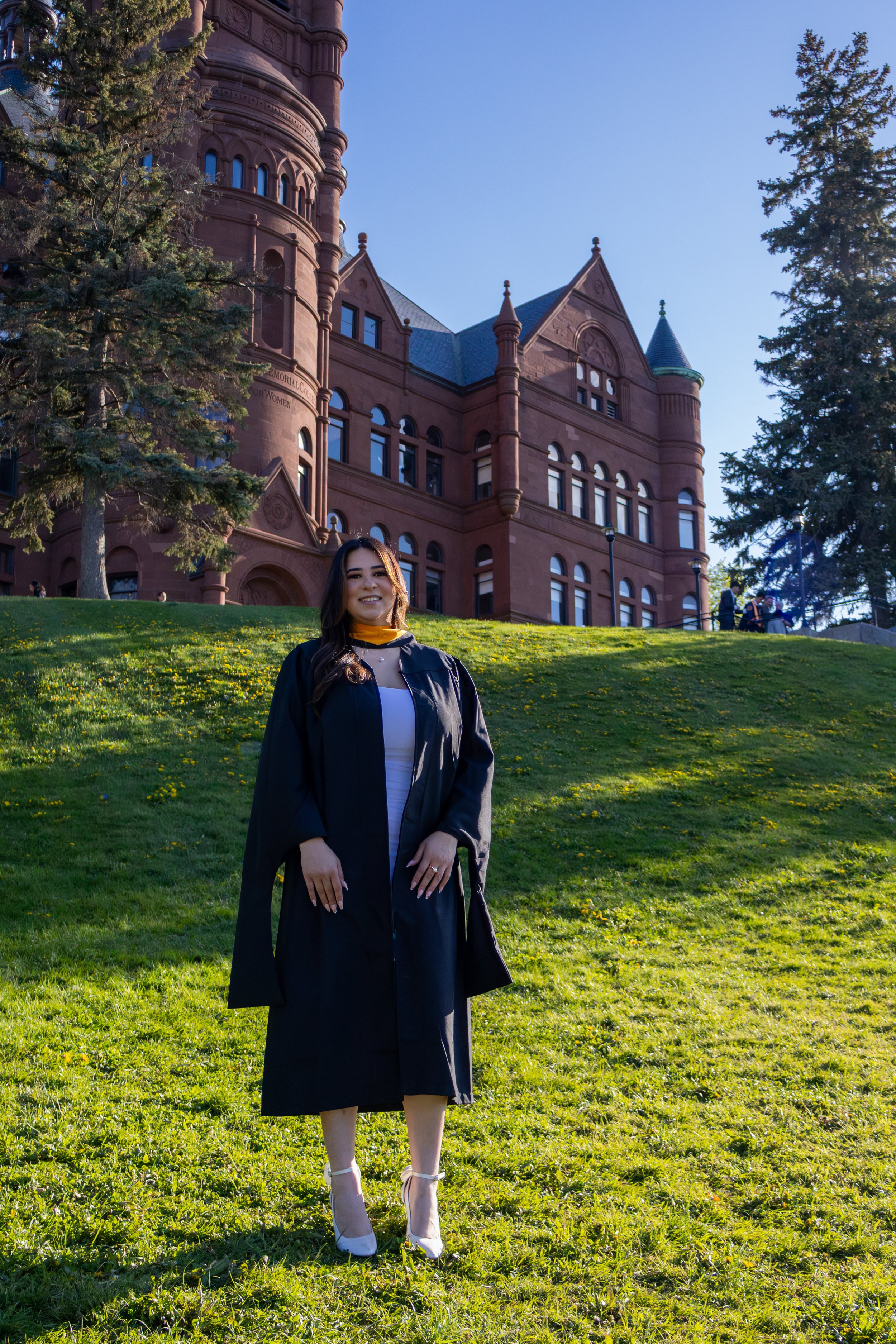 A young woman in a black graduation gown and white heels standing on a grassy hill in front of a large historic red brick building with towers, windows, and turrets. The sky is clear and the scene is illuminated by sunlight.