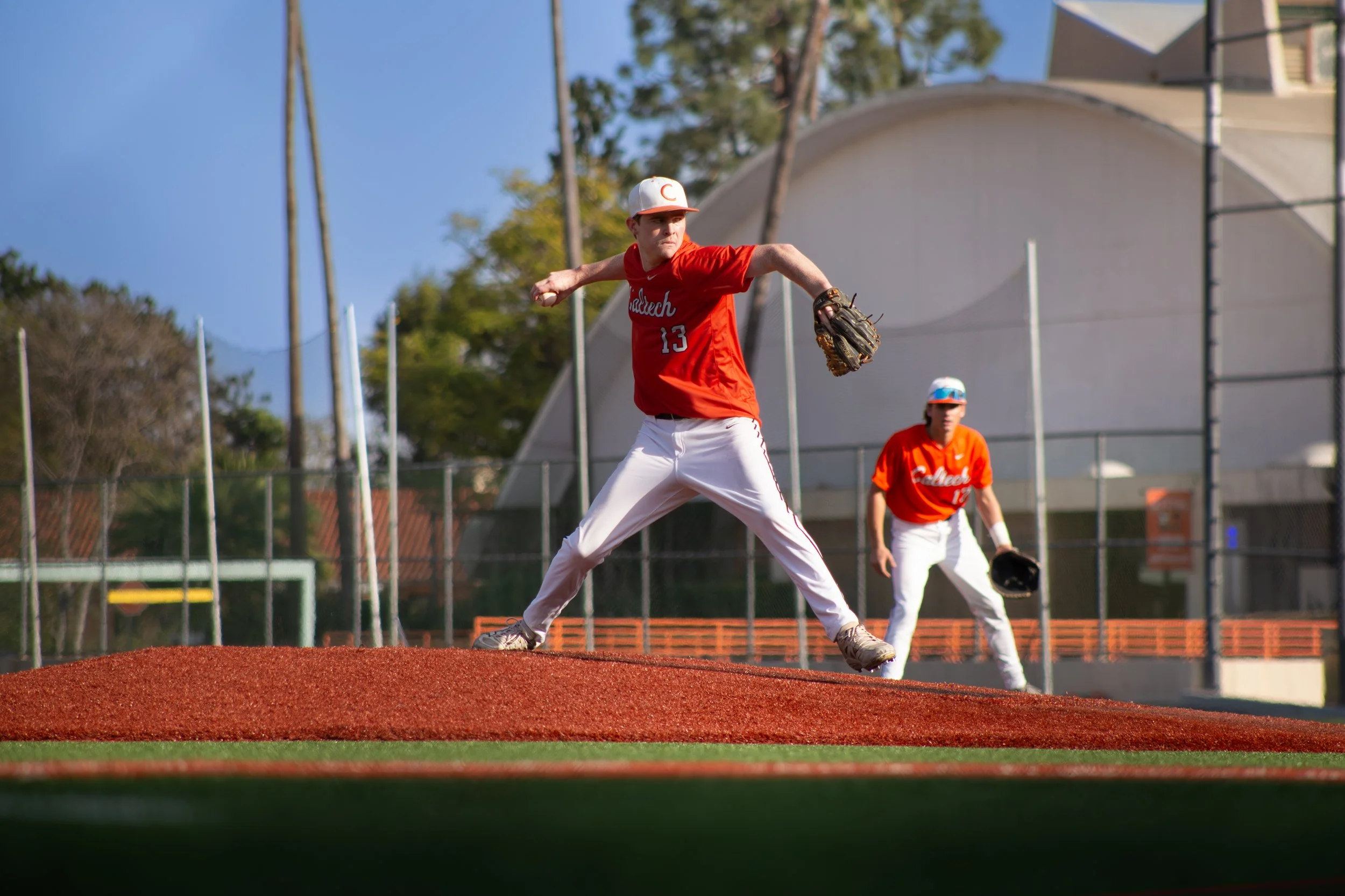 A baseball player in a red and white uniform throws a pitch on a baseball field, with another player behind him and trees in the background.