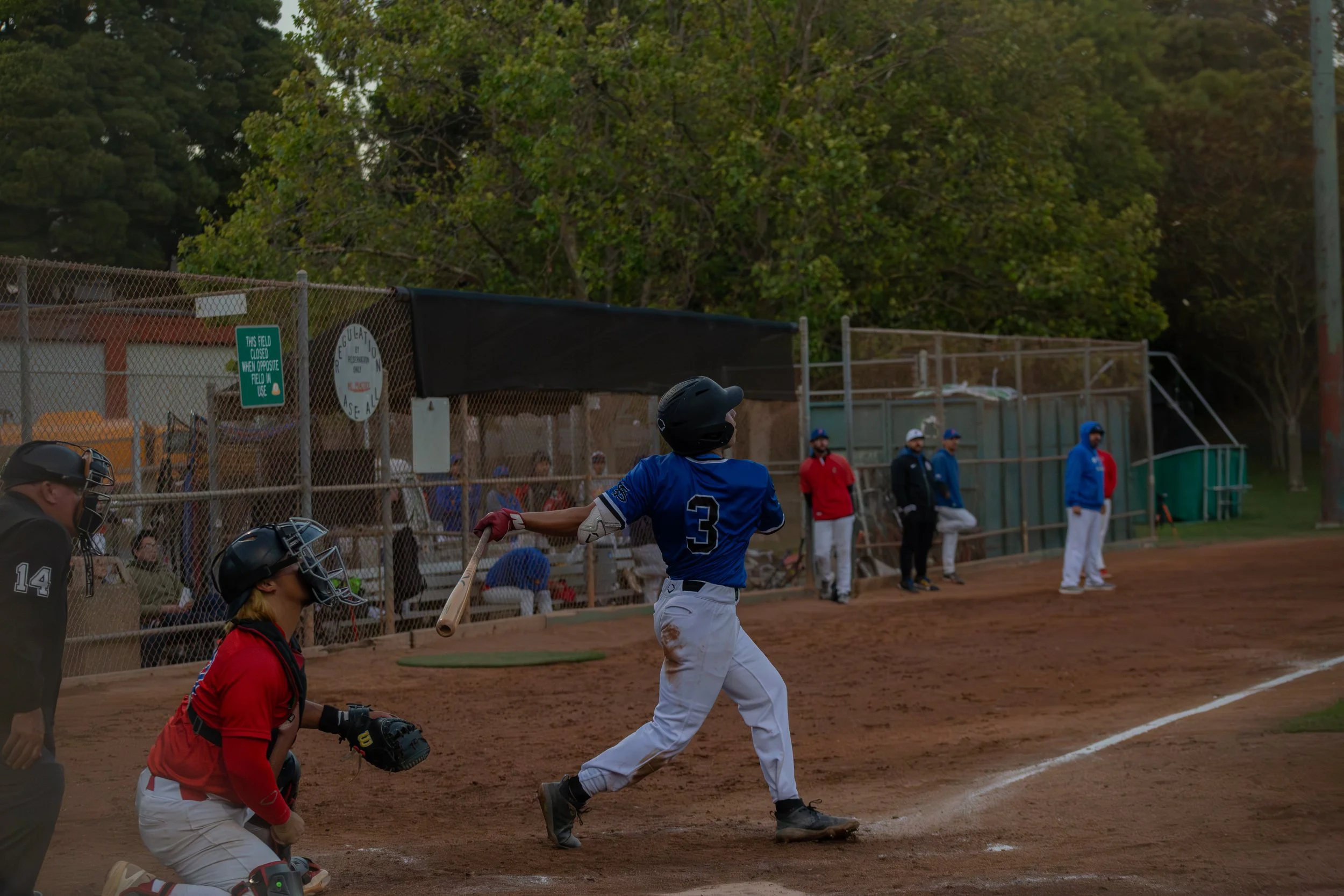 A baseball player in a white uniform and blue jersey, wearing a helmet, is swinging at a pitched ball at a baseball field during a game. An umpire and other players are watching nearby. The scene is outdoors with trees and a fence in the background.