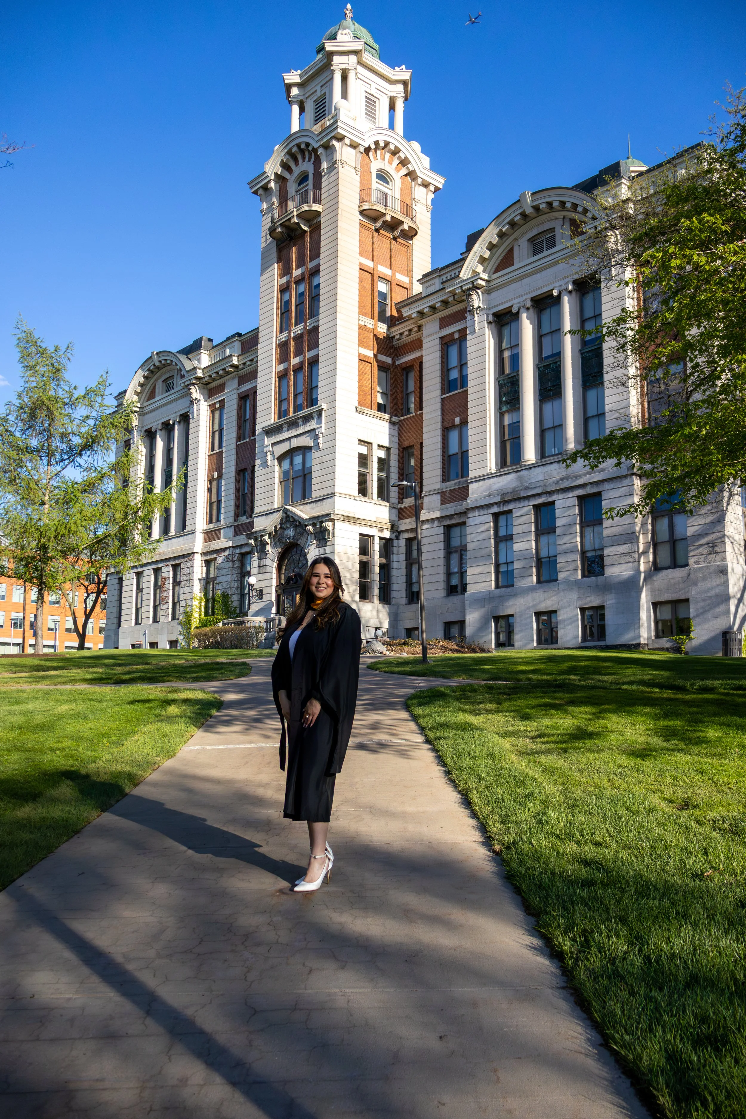 A woman in a black graduation gown and white heels standing on a sidewalk in front of a historic building with a tall clock tower. The building has white and red brick architecture, and there are trees and a clear blue sky in the background.