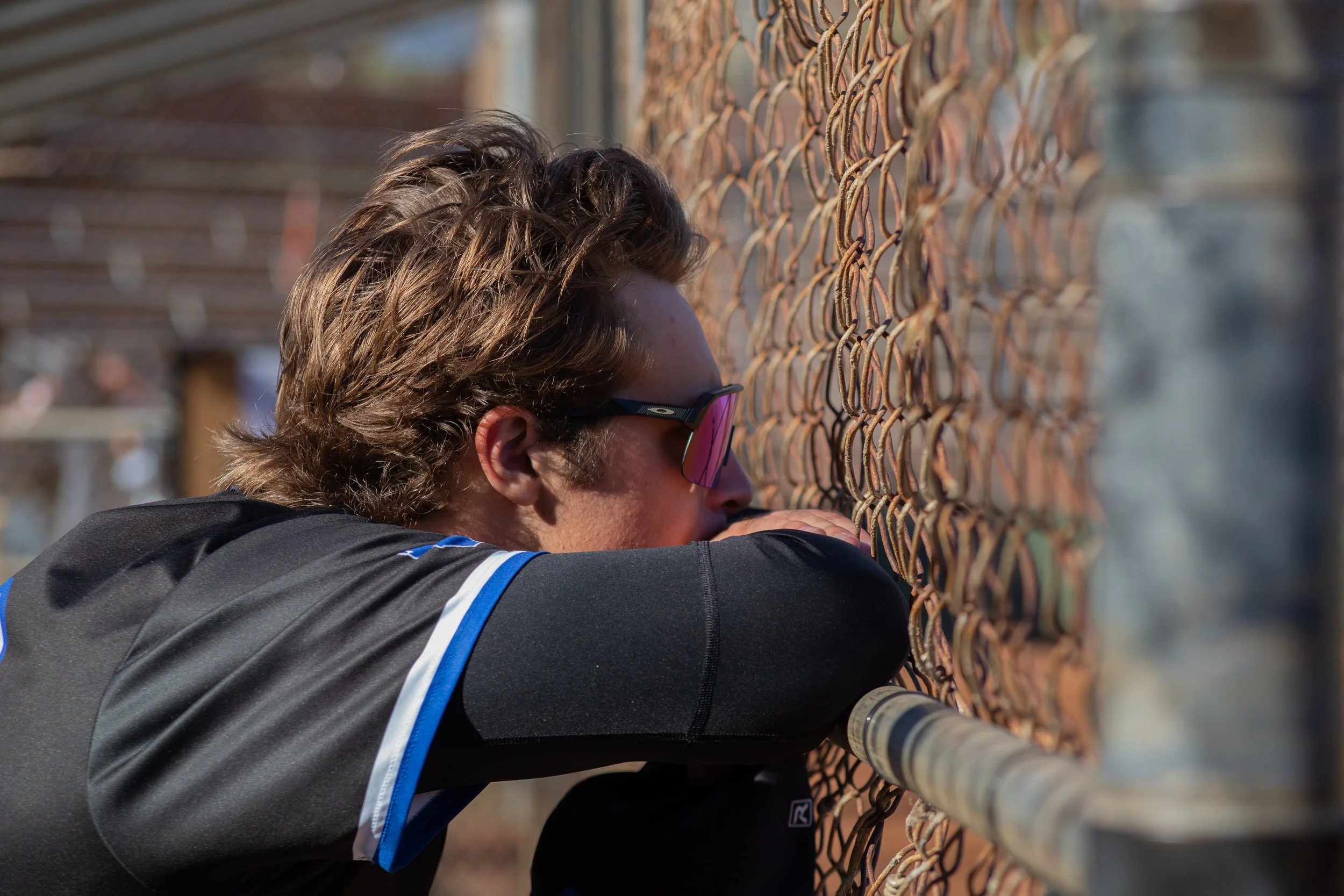 A person wearing glasses leaning on a chain-link fence with a thoughtful or contemplative expression.