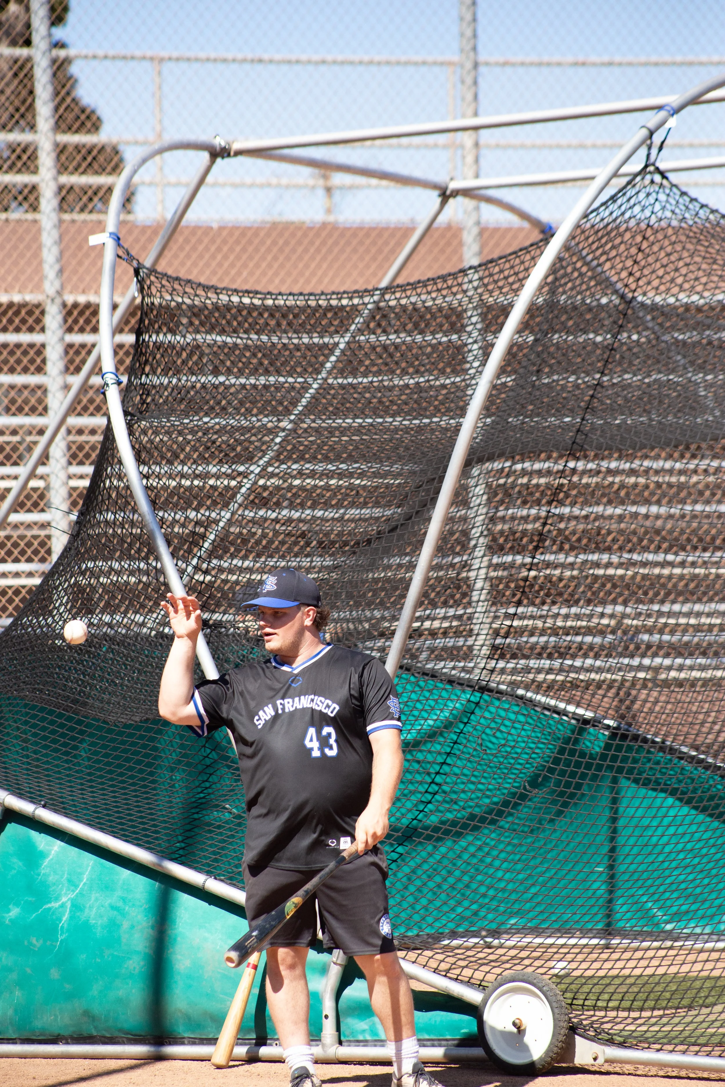 A baseball player in a black San Francisco jersey, number 43, stands near batting cage equipment, holding a bat, with a baseball in the air nearby, on a sunny day at a baseball field.