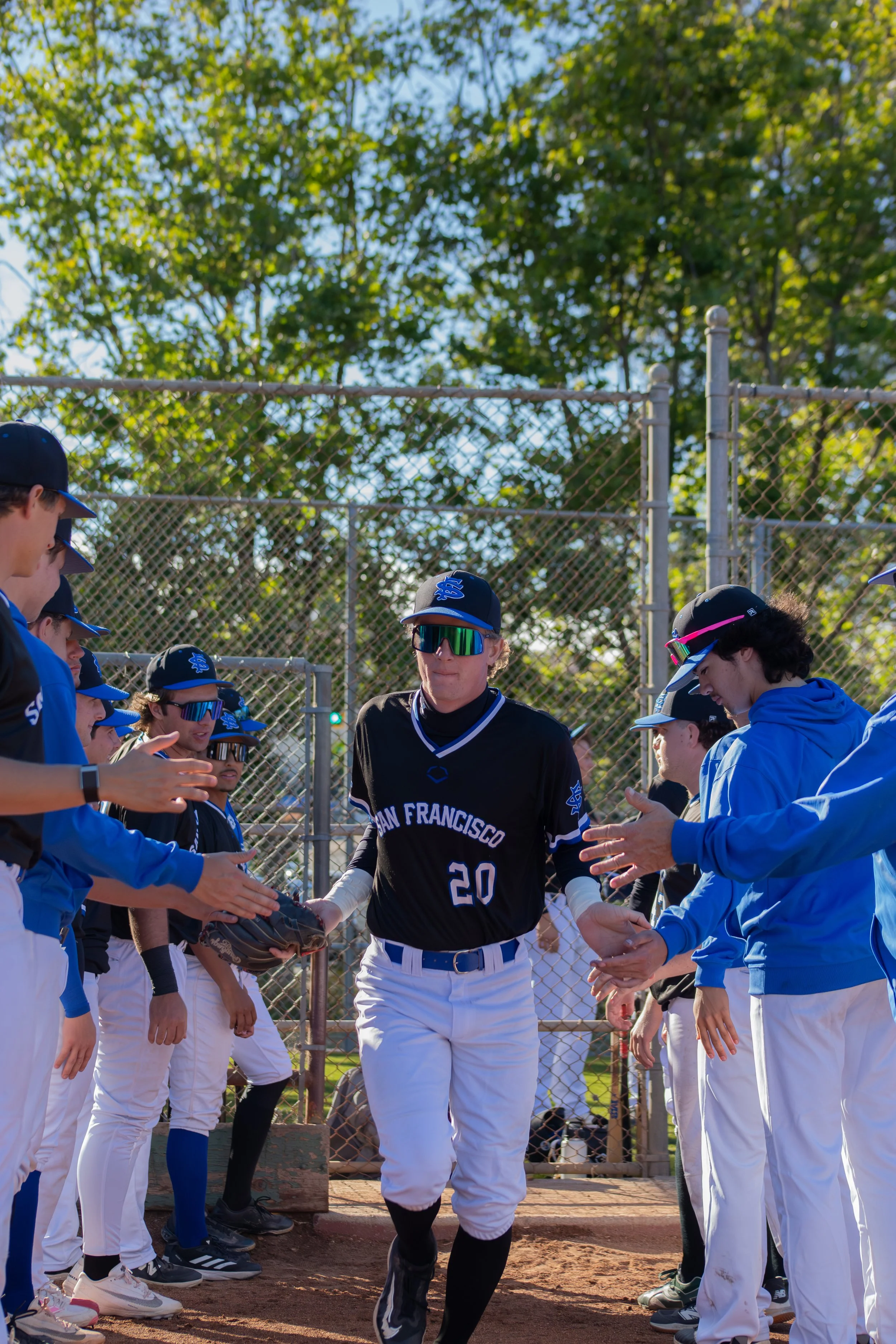 A baseball player in a black uniform with blue and white accents, gloves, and a helmet, walking through a line of teammates on a baseball field with a chain-link fence and green trees in the background.
