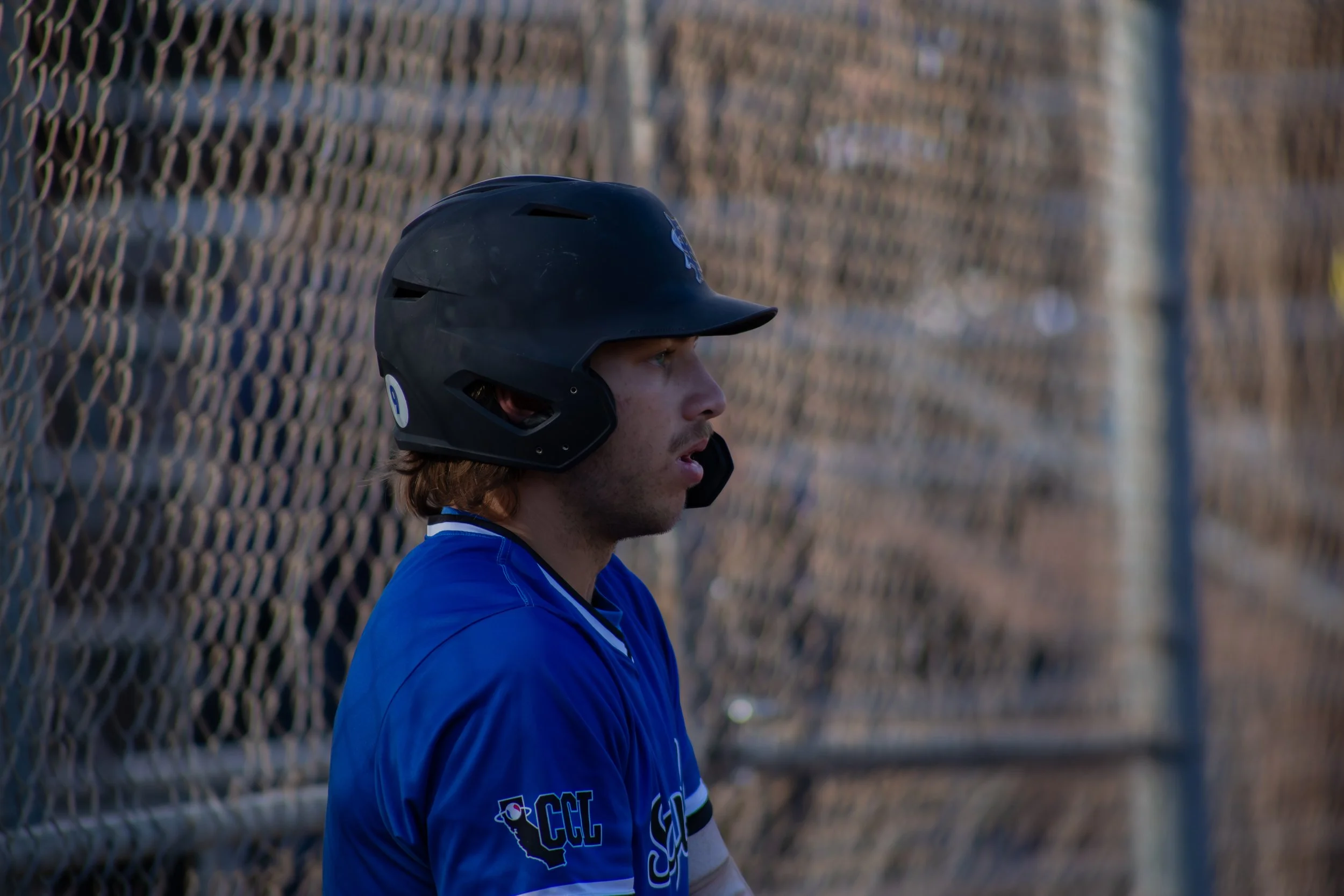 A baseball player in a blue jersey and black helmet standing near a chain-link fence.