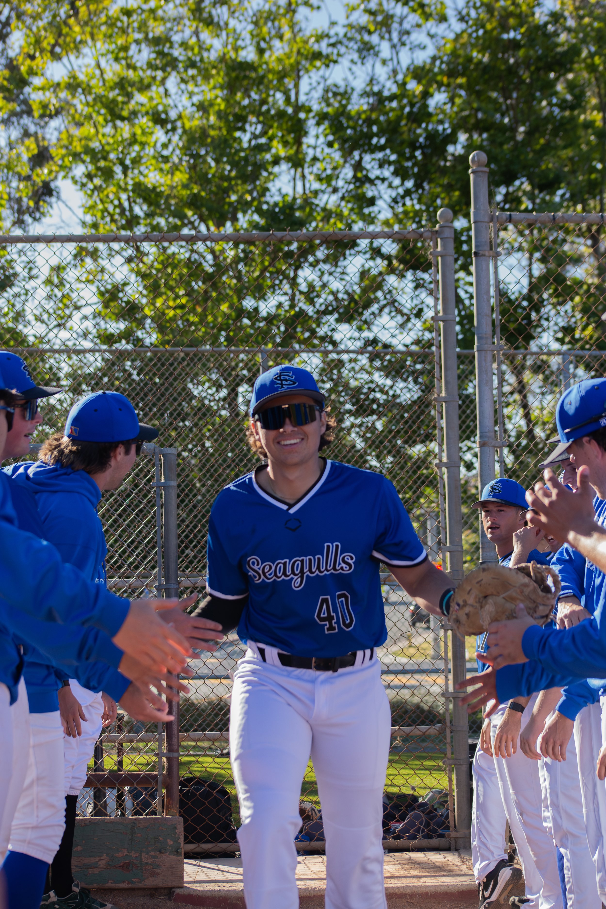 Baseball players in blue jerseys and white pants standing in a line on a baseball field, with one player in the center holding a glove, near a chain-link fence and trees in the background.
