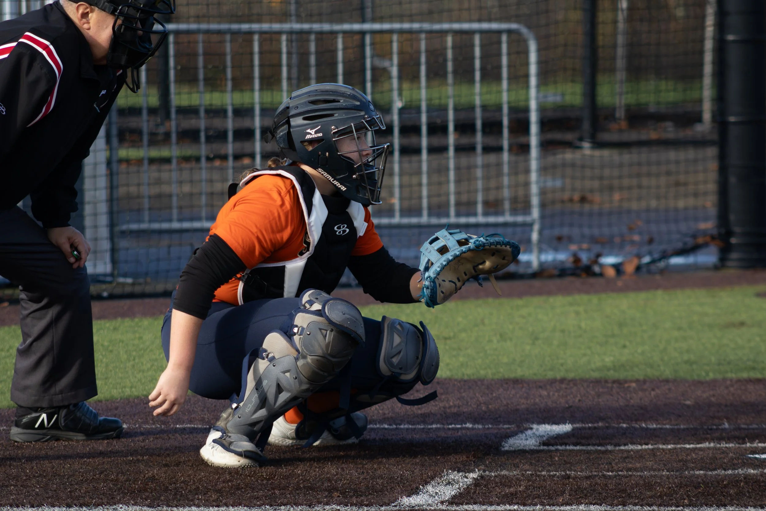 A young female baseball catcher dressed in orange, black, and gray gear squatting behind home plate on a baseball field, with a person in black coach gear leaning in talking to her.