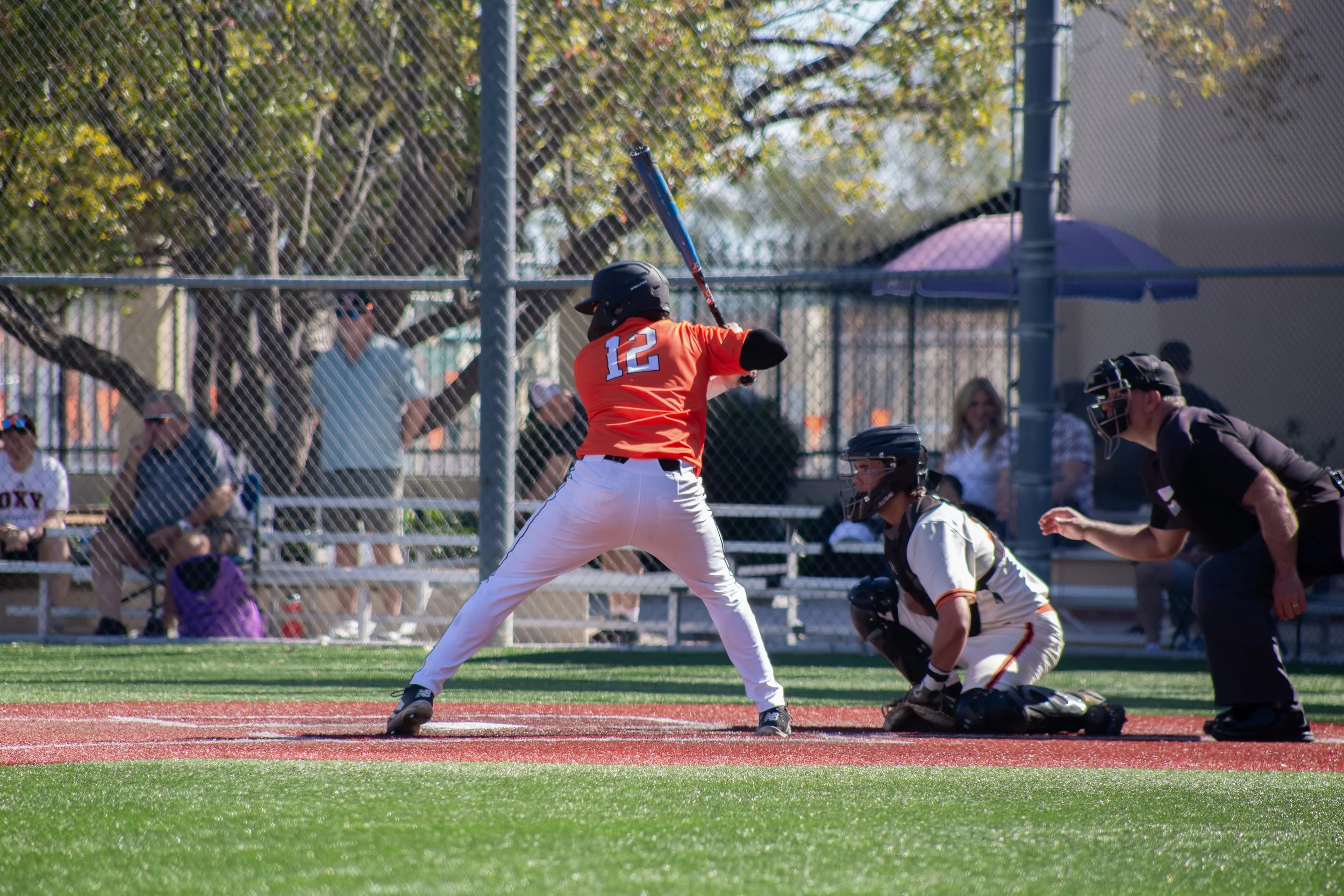 Young baseball player in an orange jersey with the number 12, swinging a bat at a pitch during a game. Spectators watch behind a chain-link fence in the background.