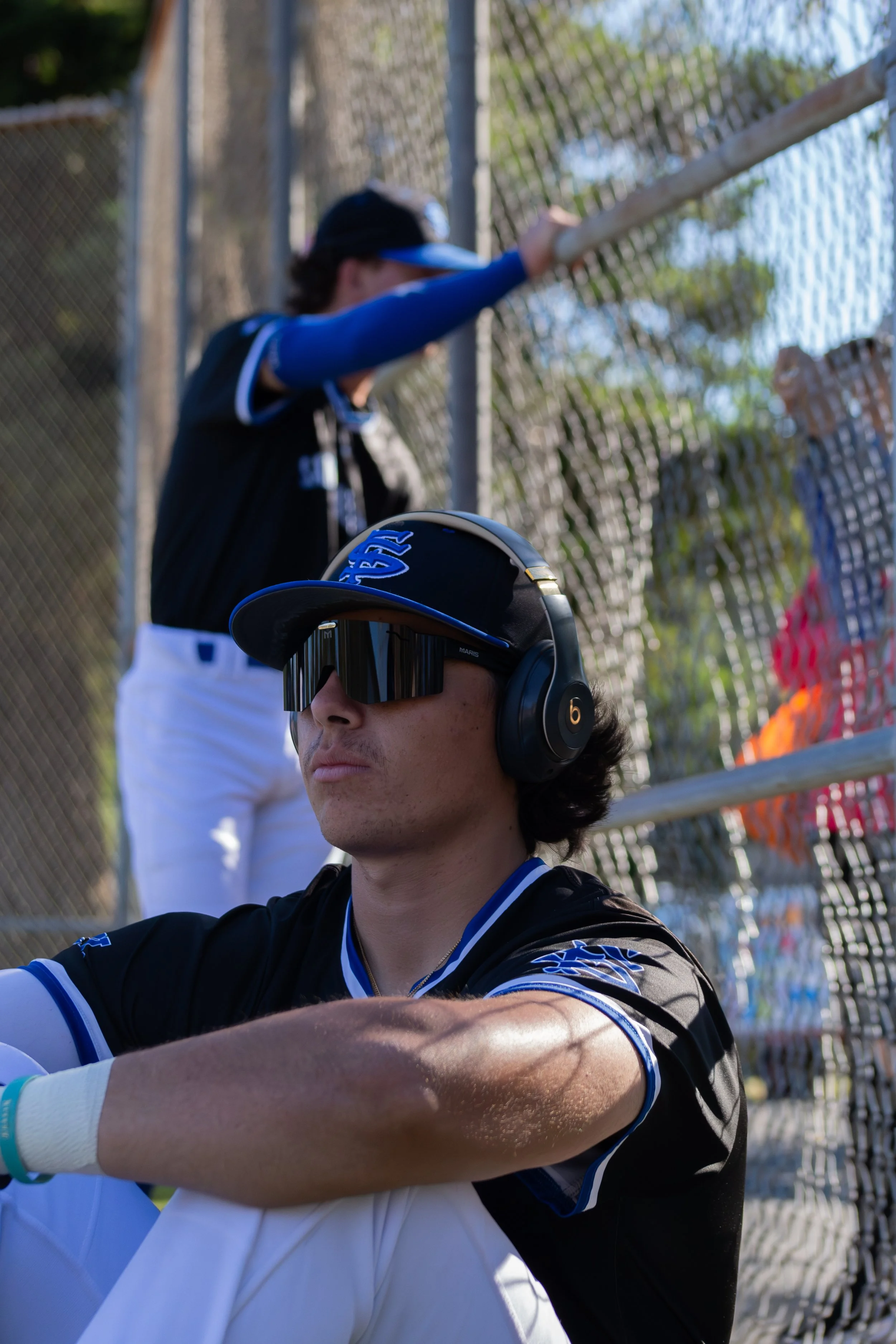 A young man wearing black and blue sports attire, large black sports headphones, and sunglasses is sitting with his arms crossed. There is a woman in sports attire and a cap standing behind him near a chain-link fence, with trees and a blue sky in th