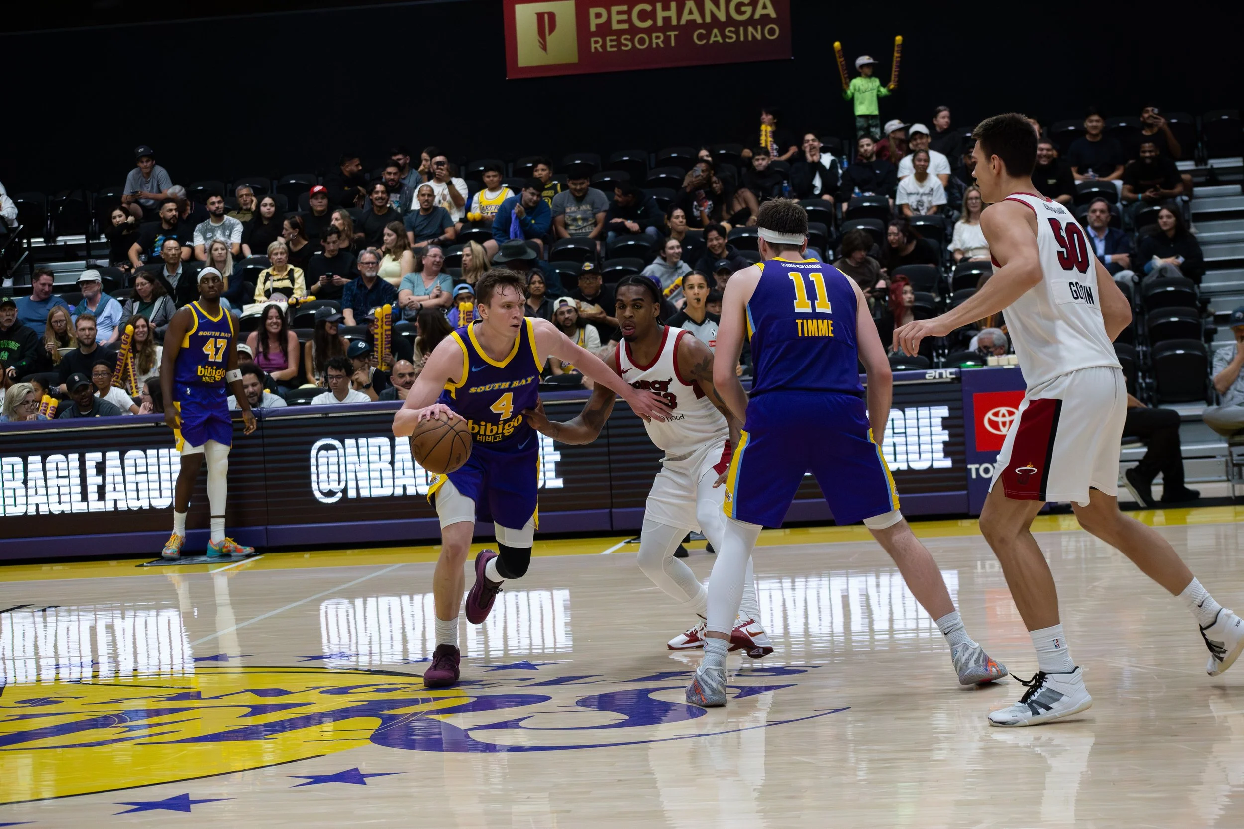 A basketball game with players from South Bay and the opposing team in white jerseys, with a man in a white and red jersey holding the basketball and looking to pass, while other players and spectators watch from the stands.