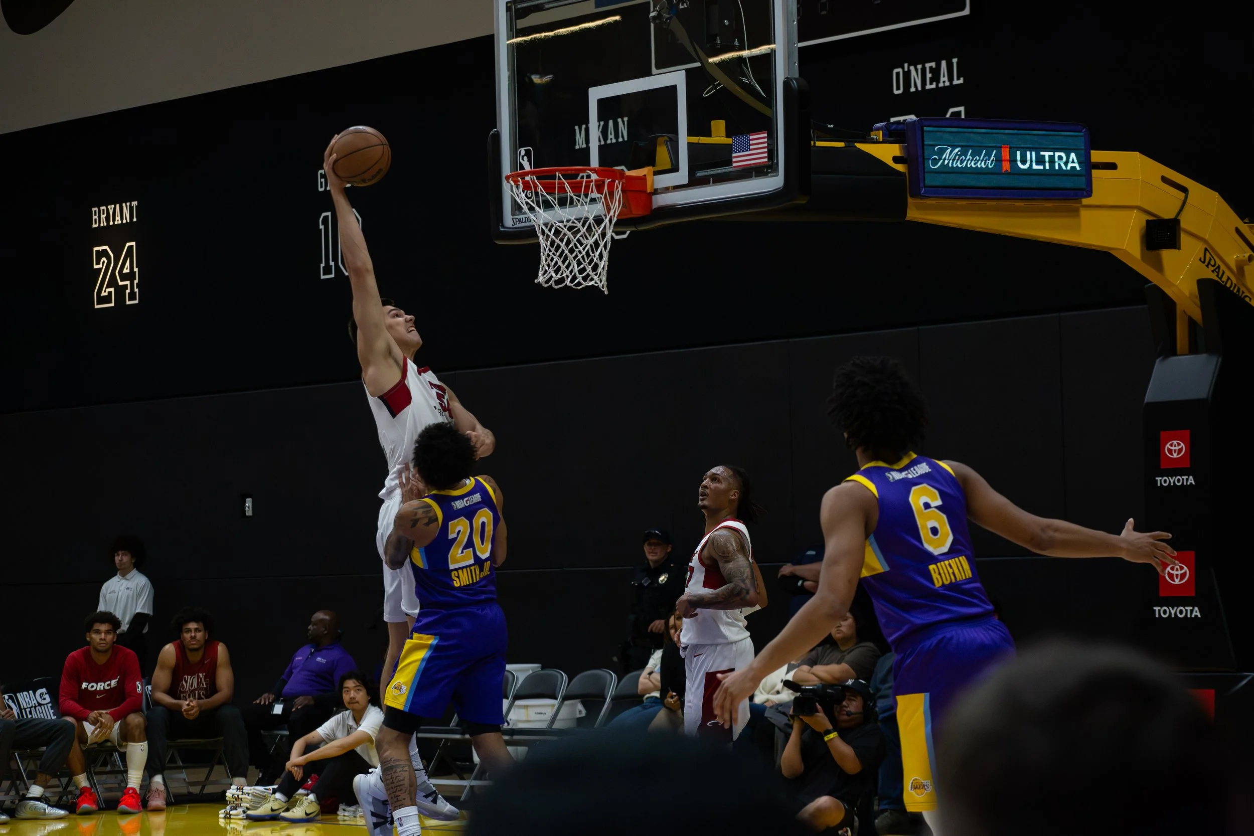 A basketball player in white jersey jumping to score near the hoop with defenders in blue jerseys nearby, and spectators watching from the sidelines.