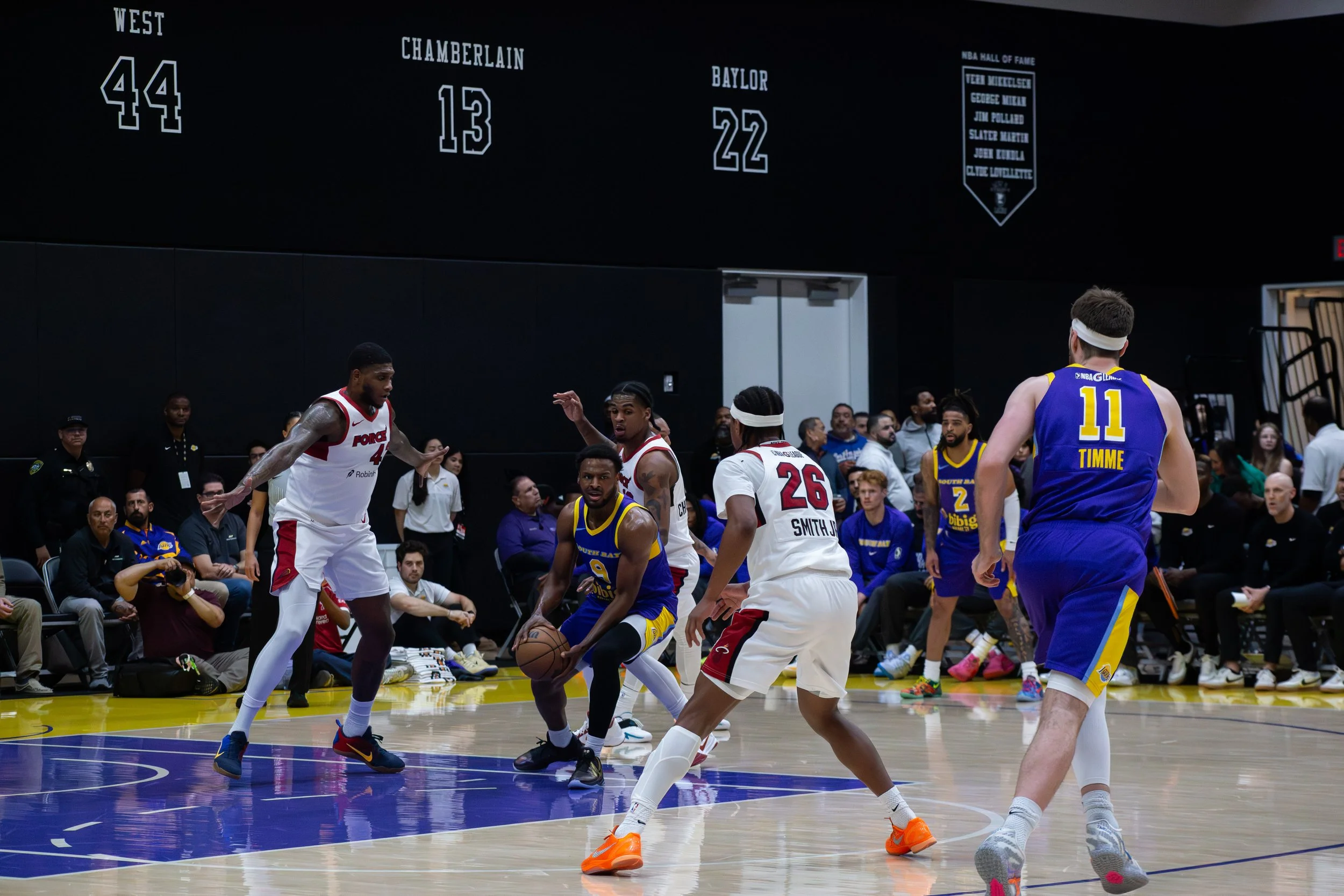 Basketball game with players from two teams, one in white jerseys and the other in blue and yellow jerseys, on a court with spectators and officials watching.