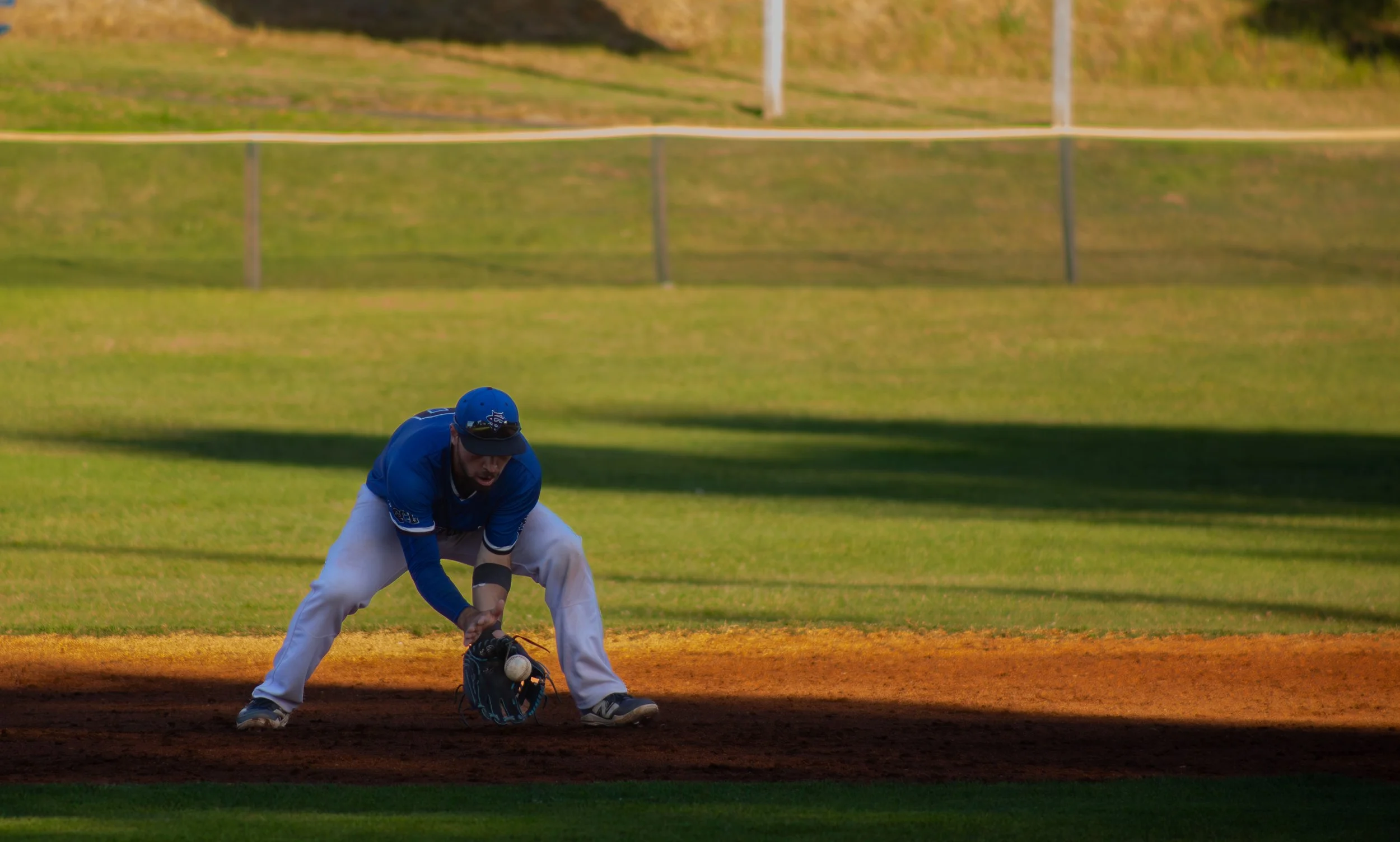 A baseball player in a blue uniform and cap is fielding a ground ball on a baseball field, positioned near the infield dirt with green grass in the background.