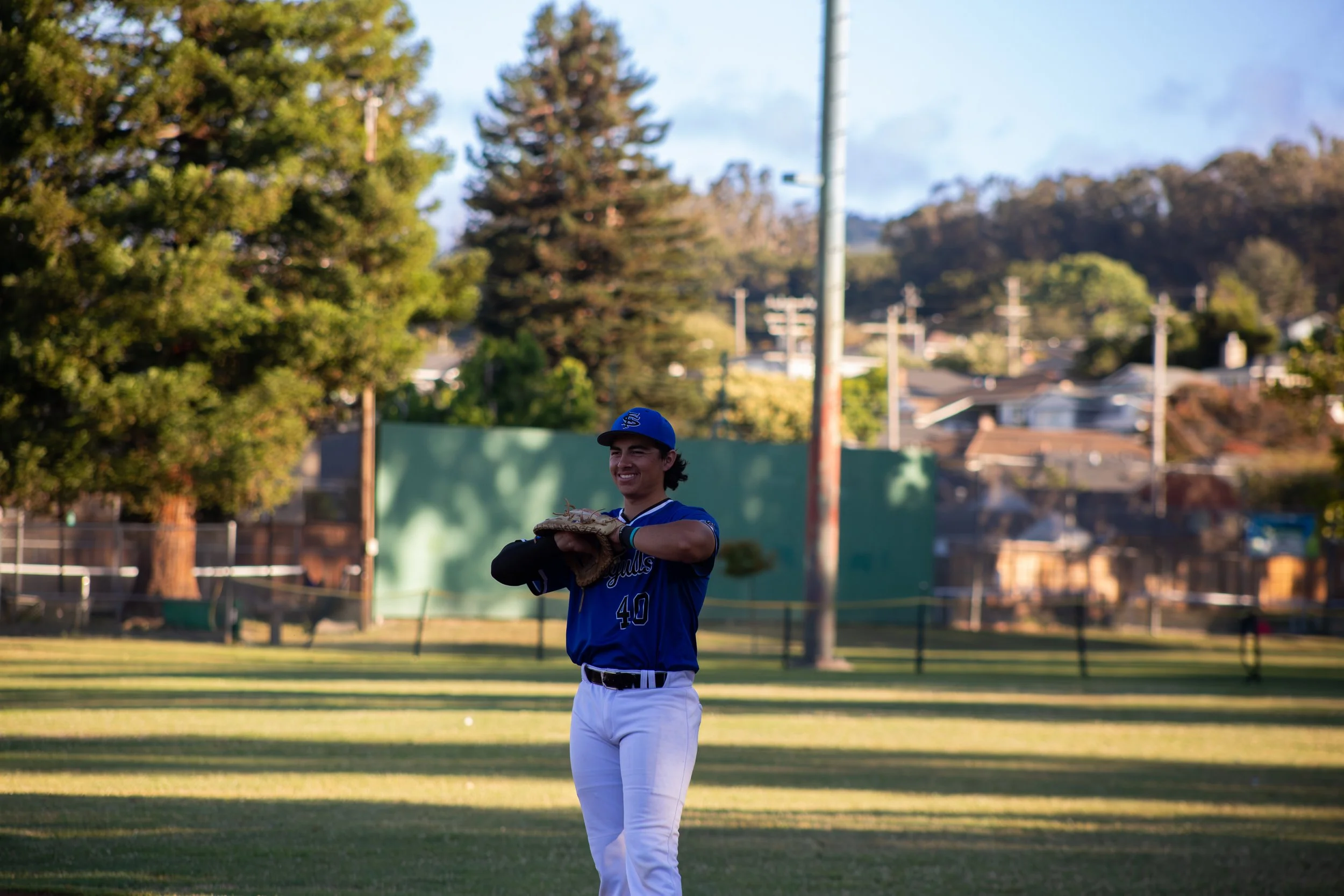 A young baseball player in a blue jersey and white pants standing on a baseball field, smiling, holding a glove, with trees and utility poles in the background during the daytime.