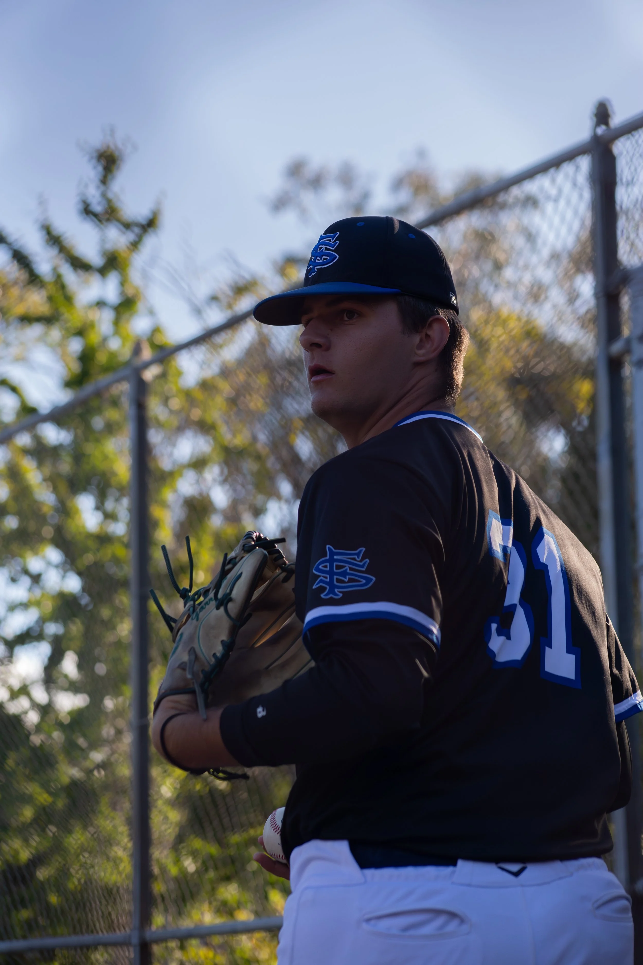 A young man wearing a black baseball cap and a black and blue baseball jersey stands outdoors near a chain-link fence, holding a baseball glove on his left hand and a baseball in his right hand, with trees and a blue sky in the background.