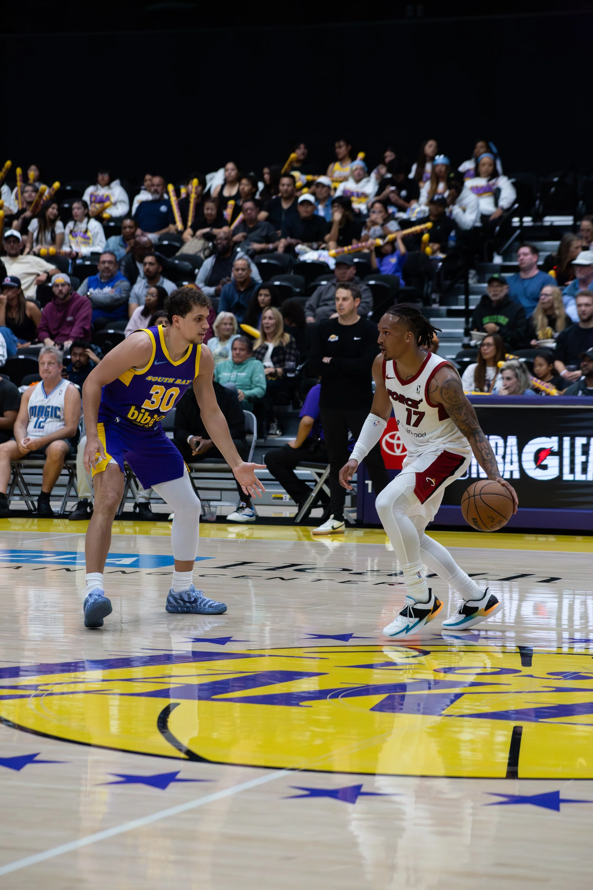 Two basketball players, one in a blue and yellow jersey and the other in a white and red jersey, are playing during a game. The crowd in the background is watching, some holding yellow noise-making sticks.