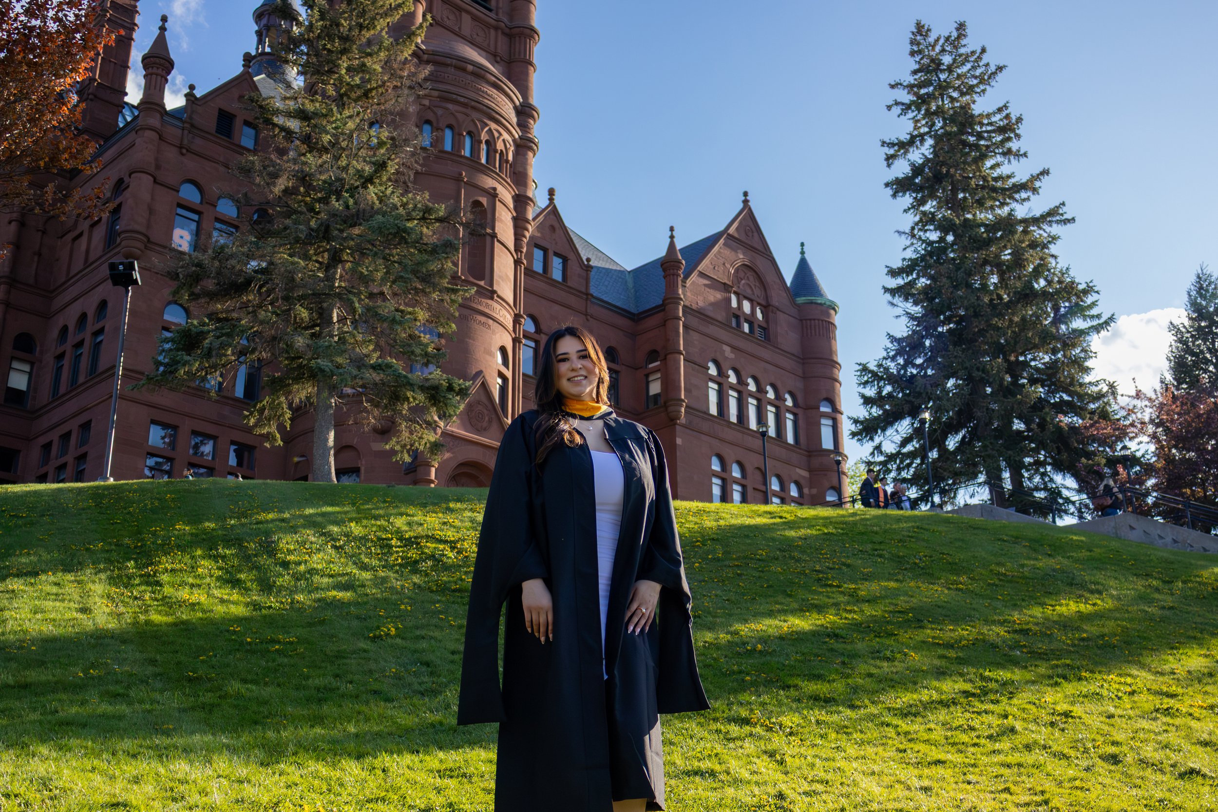 Young woman in graduation gown and cap standing on a grassy area in front of a historic red sandstone building with turreted towers, trees, and a blue sky.