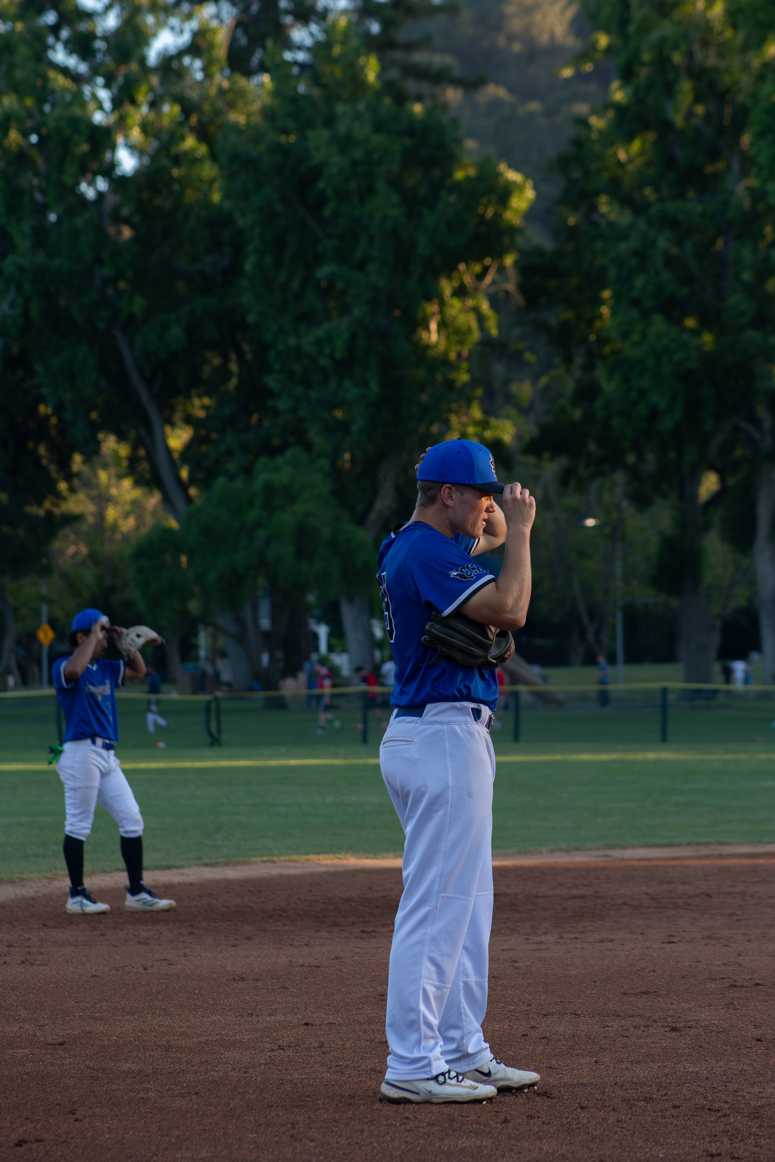 Baseball players in blue uniforms standing on the field, one adjusting his cap, during a game in a park with trees in the background.