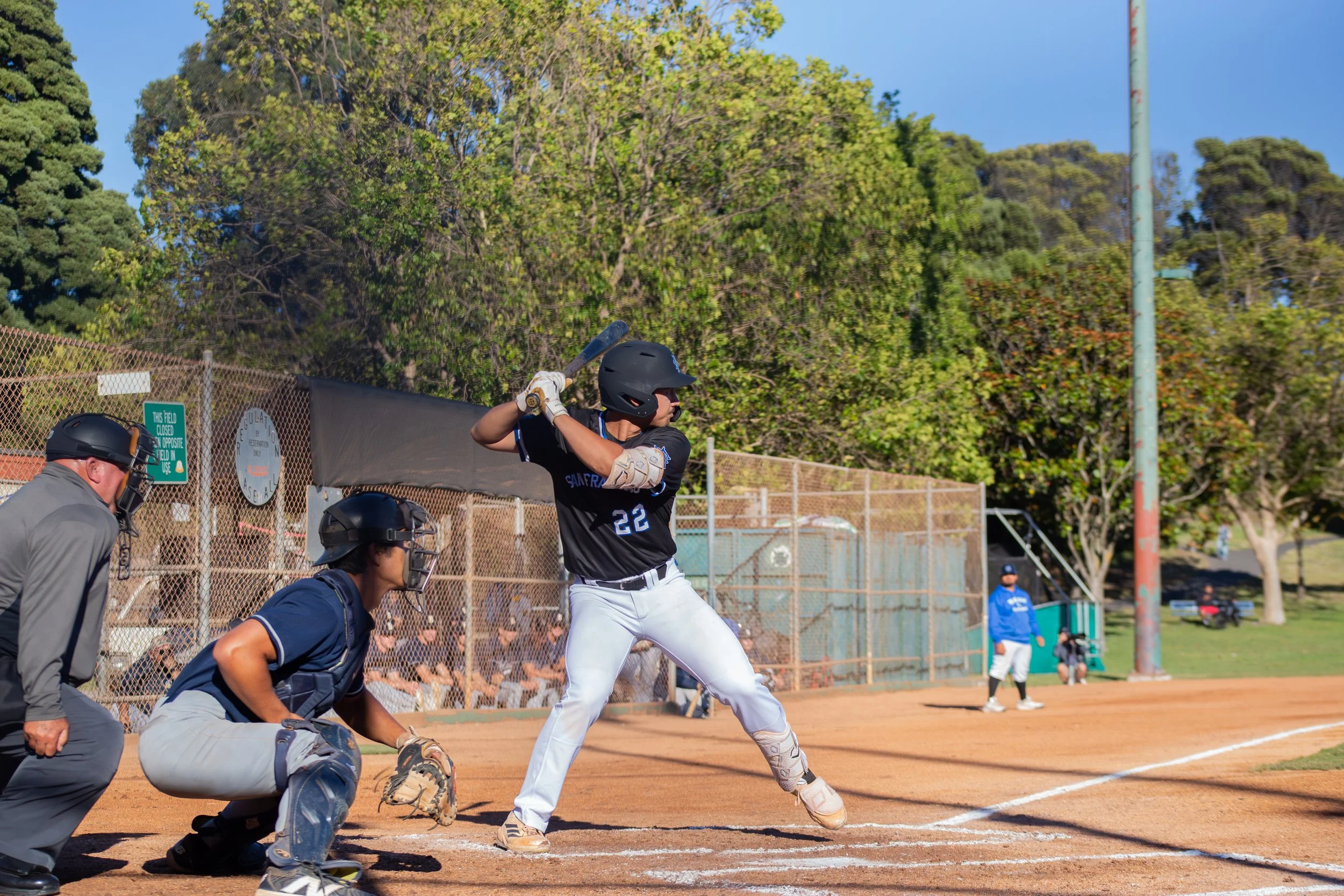 A baseball player at bat during a game, with a batter in a black jersey and white pants, surrounded by team members and coaches on a dirt field with a chain-link fence and trees in the background.