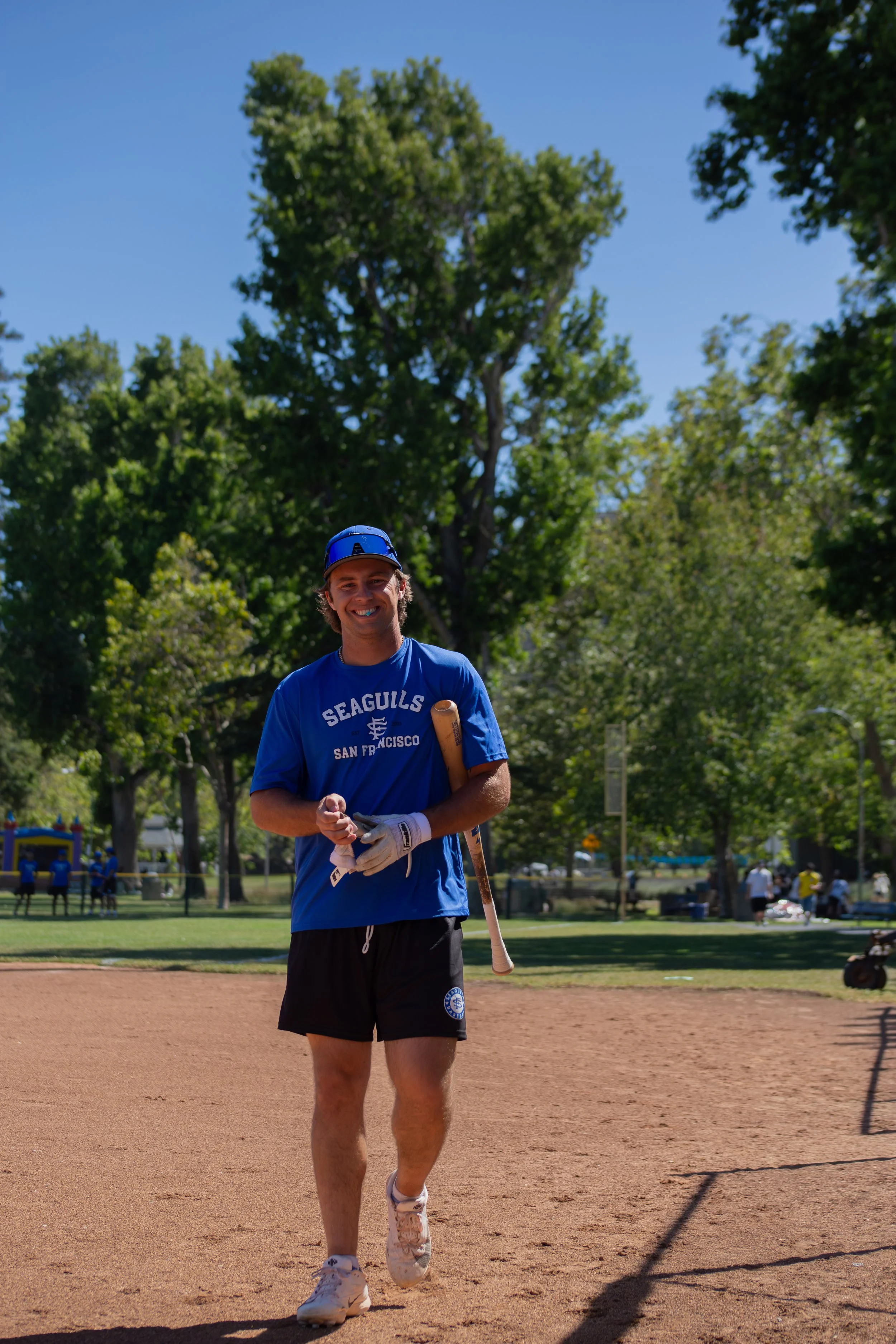 A smiling young man wearing a blue baseball cap, blue t-shirt with 'Seagulls San Francisco' printed on it, black shorts, and white sneakers, holding a baseball bat, standing on a baseball field with trees and other people in the background on a sunny