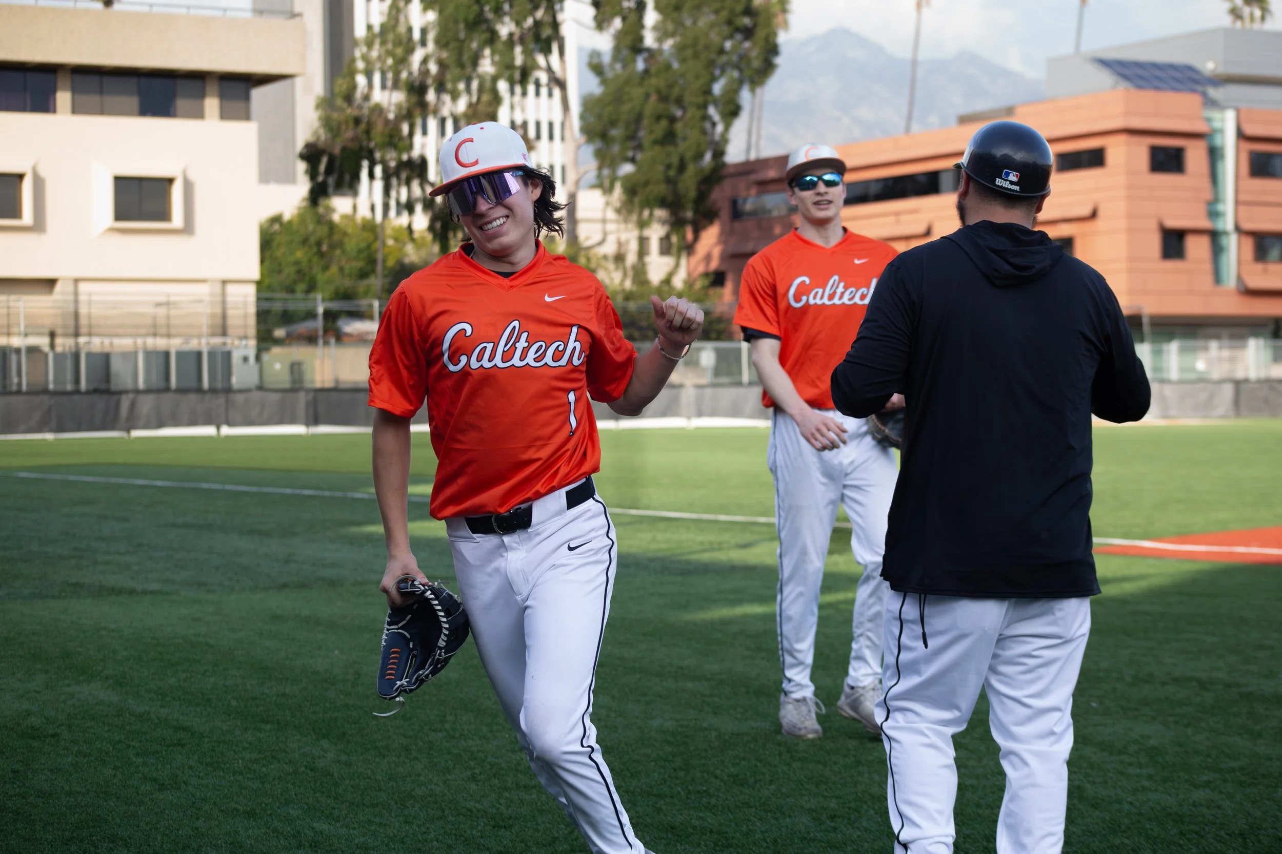 Two baseball players from Caltech wearing orange jerseys and white pants, paired with a person in black clothing and helmet, converse on a green sports field, with modern buildings and trees in the background.