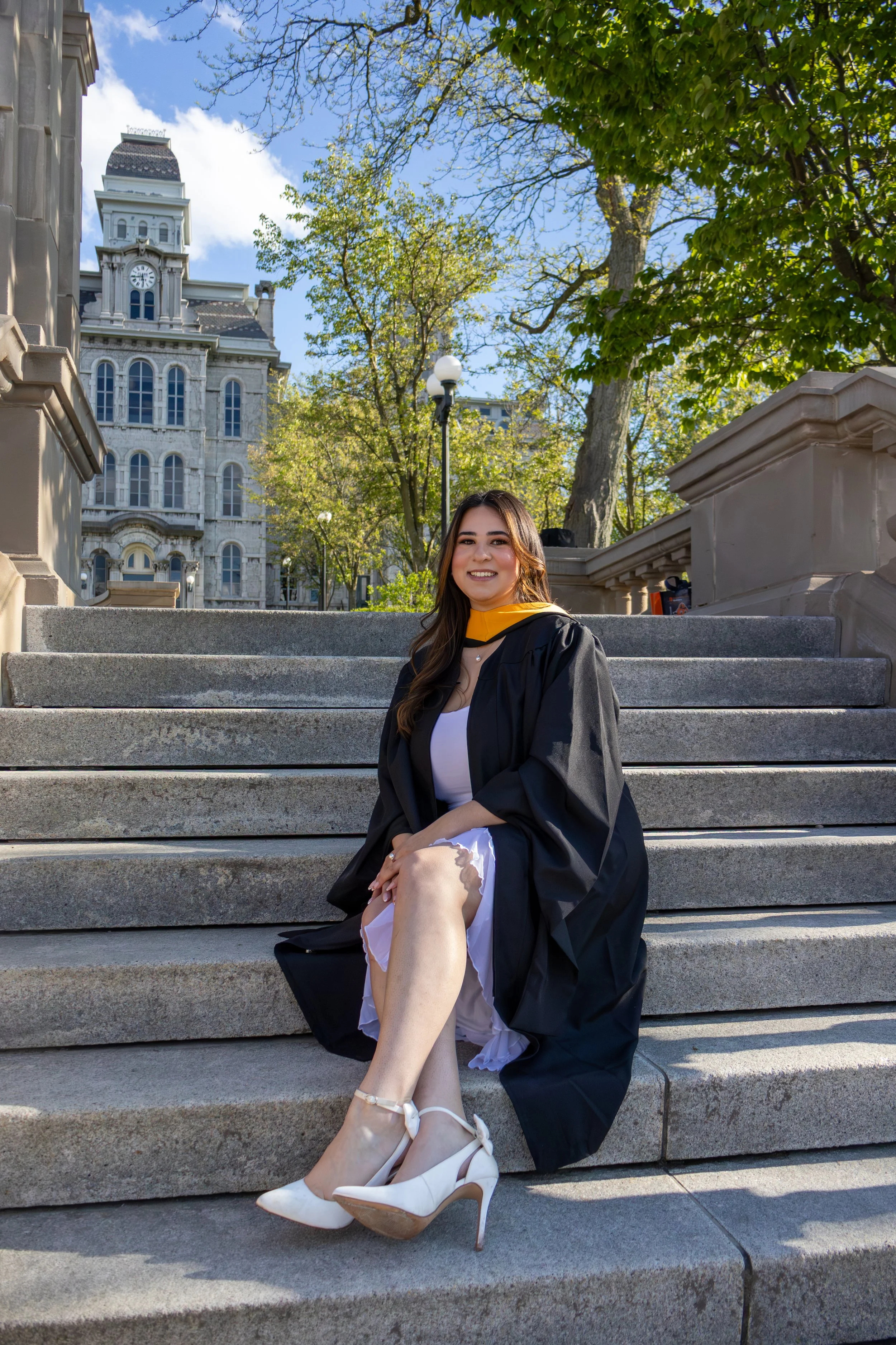 A young woman in a graduation gown and cap sitting on steps outdoors, smiling with a historic building and trees in the background on a sunny day.