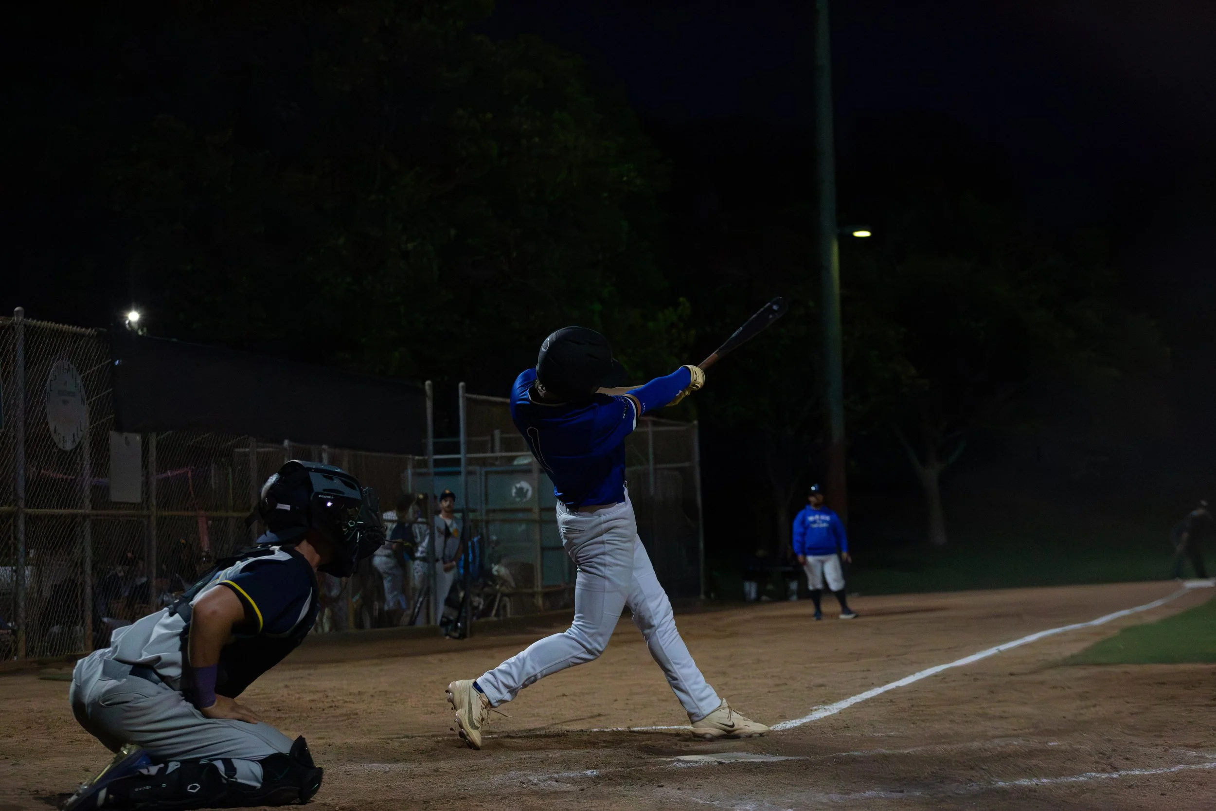 A baseball player swinging at a pitch during night game, with players and spectators in the background.