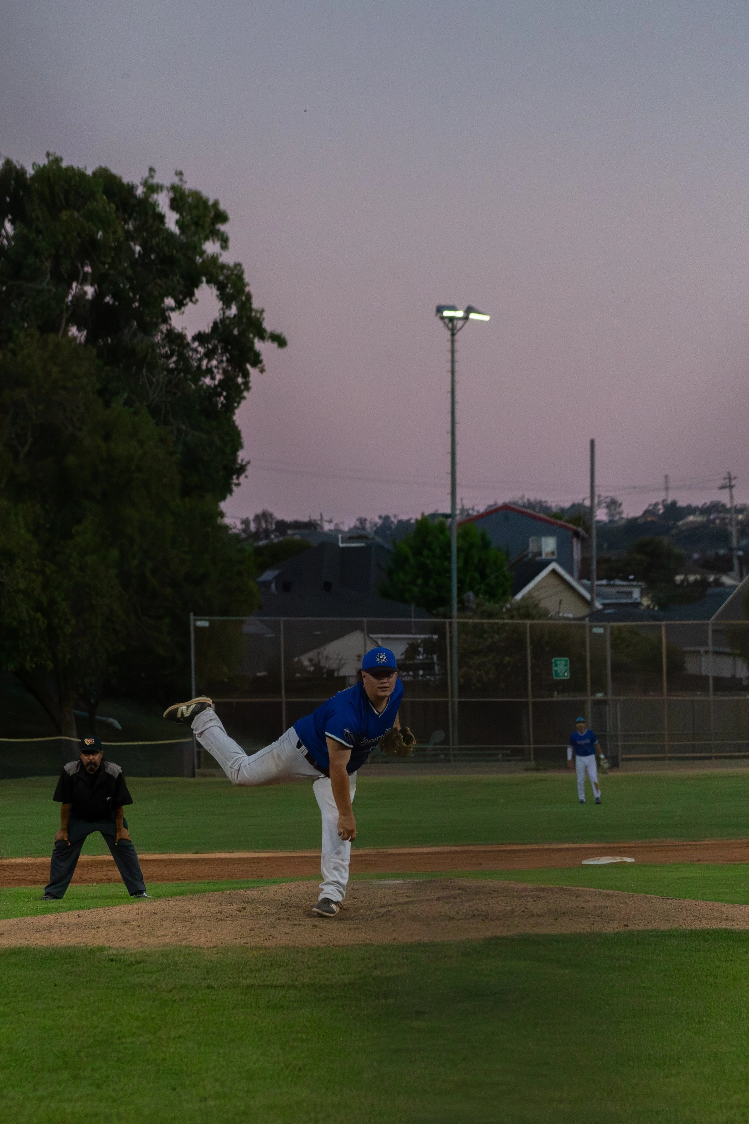A baseball pitcher in a blue uniform is in the act of pitching the ball. Other players and an umpire are visible on the field, with trees and houses in the background during early evening.