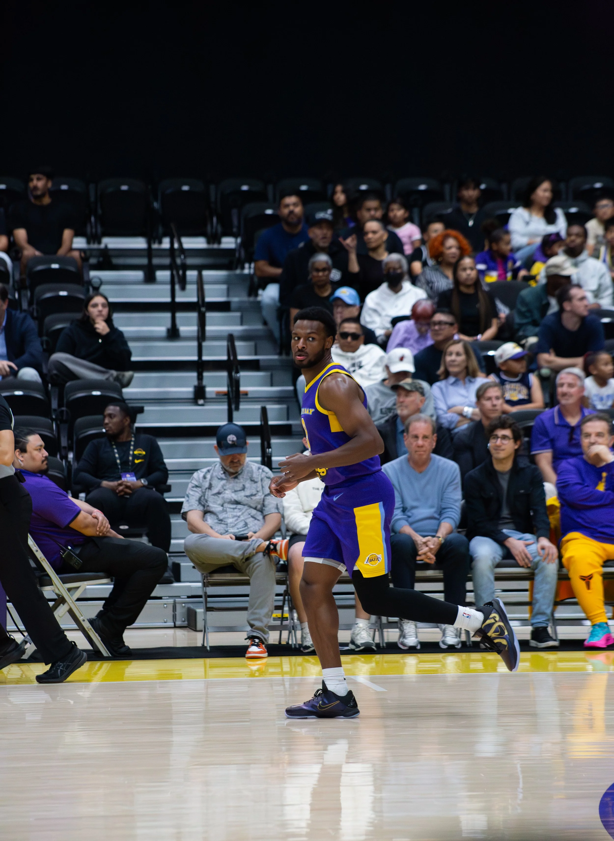 A basketball player wearing a purple and yellow Los Angeles Lakers jersey is running on the court during a game, with an audience seated in the background.