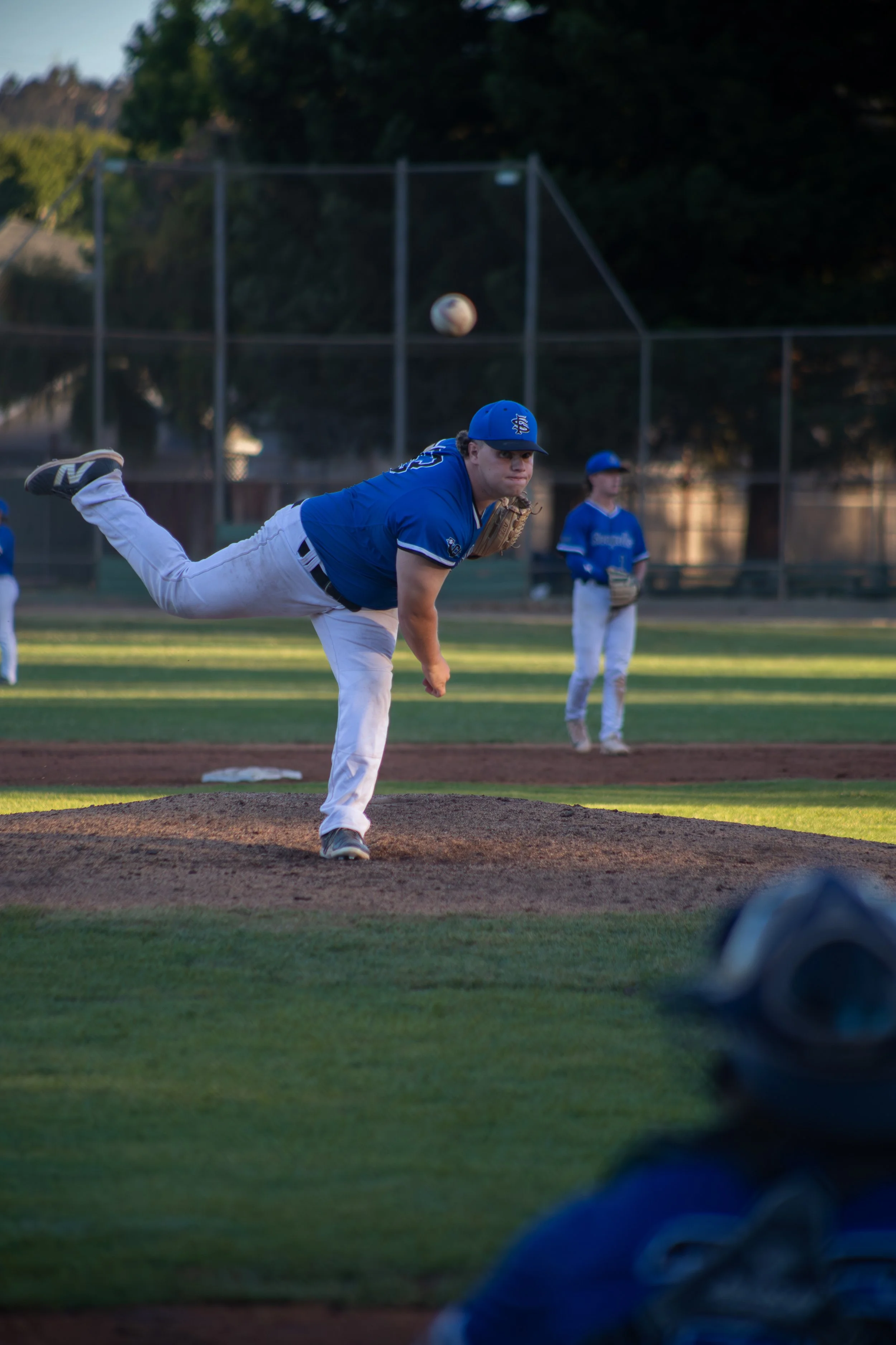 A young baseball player in a blue uniform and cap pitches a ball on a baseball field during daylight. Other players are visible in the background, and the background features a fence and trees.