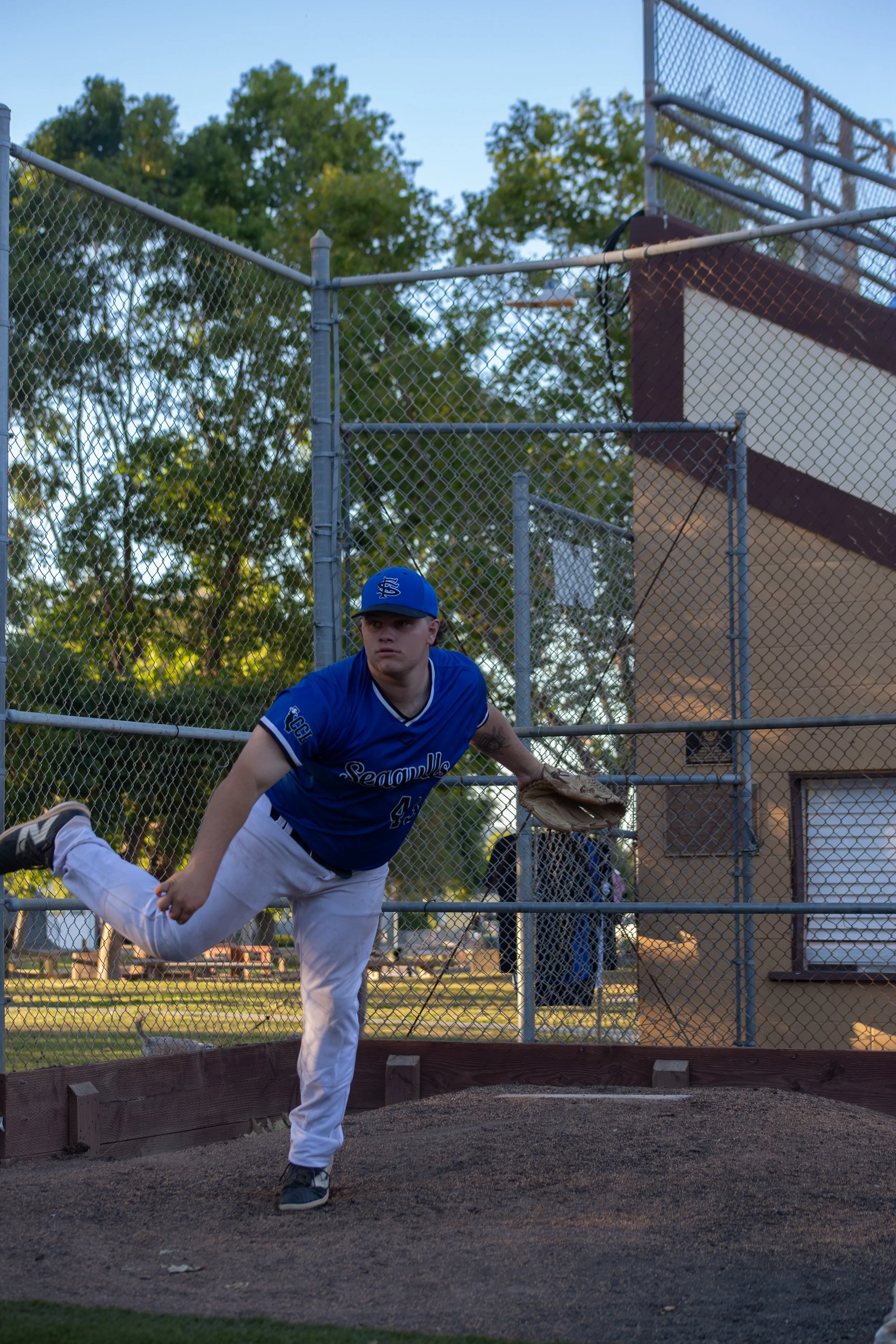 Baseball player in blue jersey throwing a pitch on a baseball field, with trees and a chain-link fence in the background.