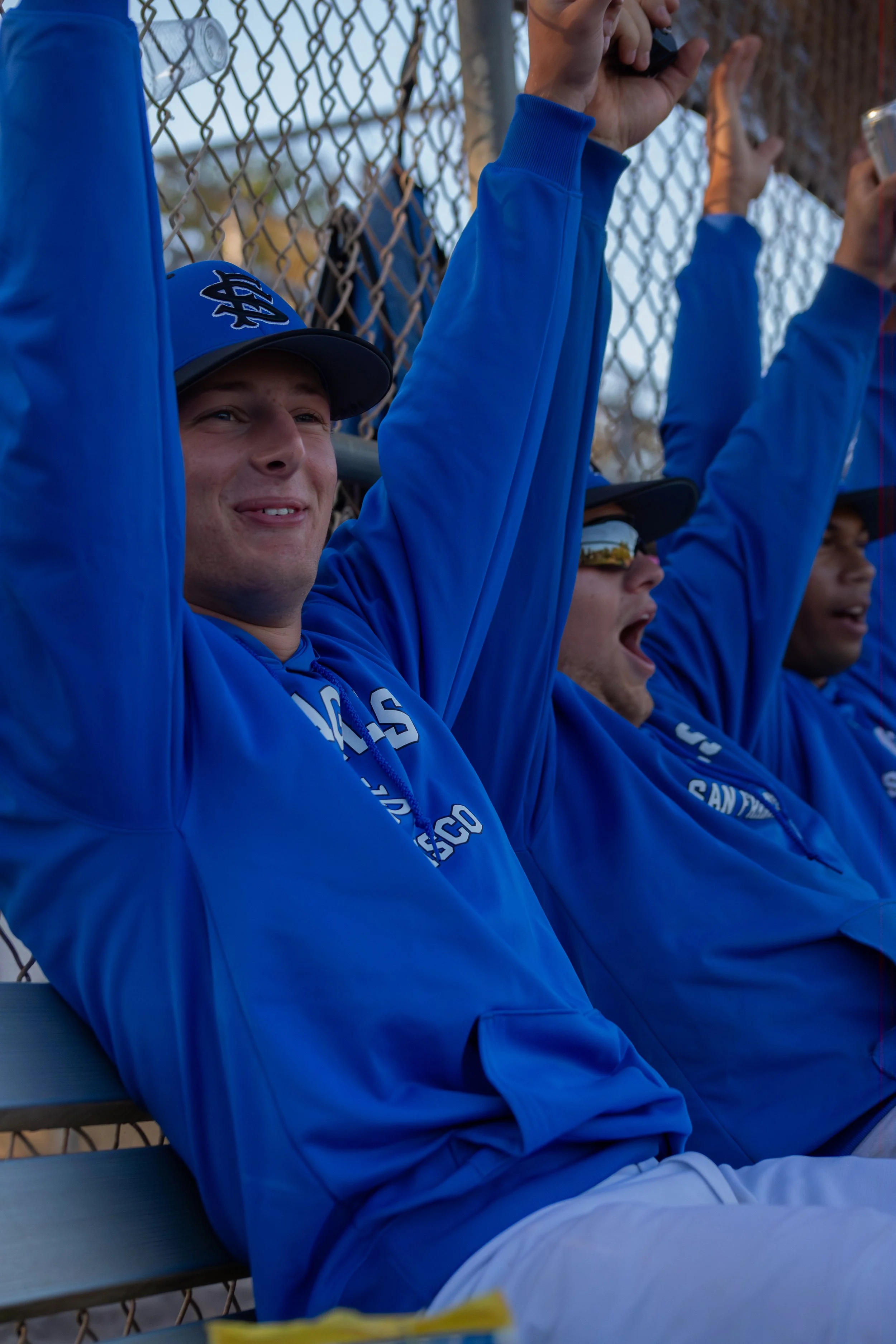 Baseball players in blue jerseys and caps sitting on a bench near a chain-link fence, some raising their hands or holding phones, smiling and cheering.