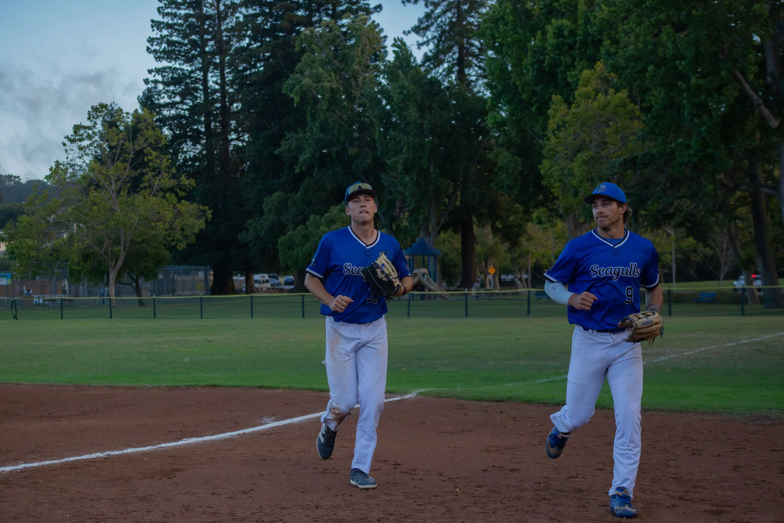 Two baseball players in blue uniforms and white pants jogging on a baseball field during evening time. They are wearing caps and gloves, with a background of trees and parked cars.