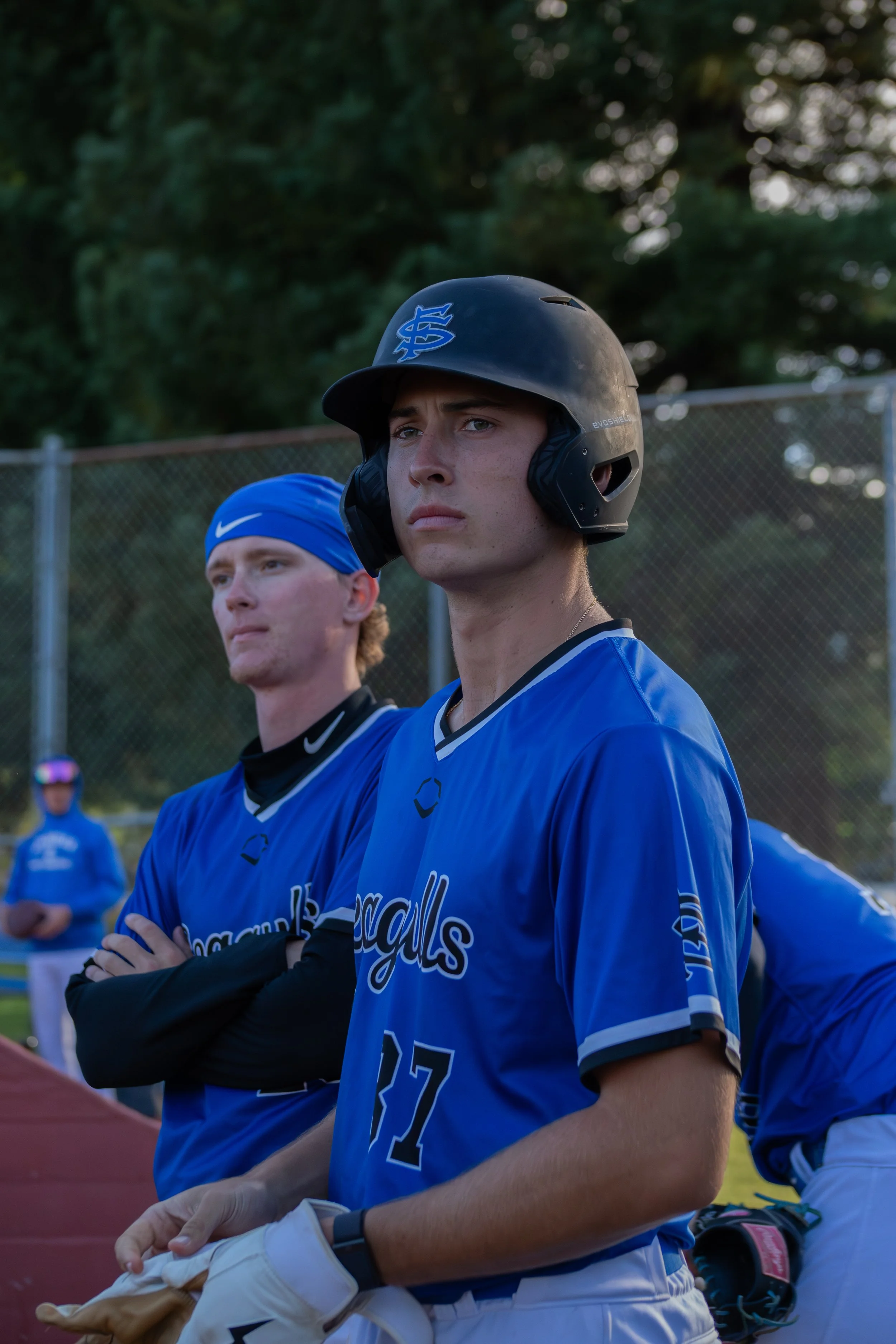 Two young male baseball players wearing blue jerseys and helmets standing on a baseball field during daytime.