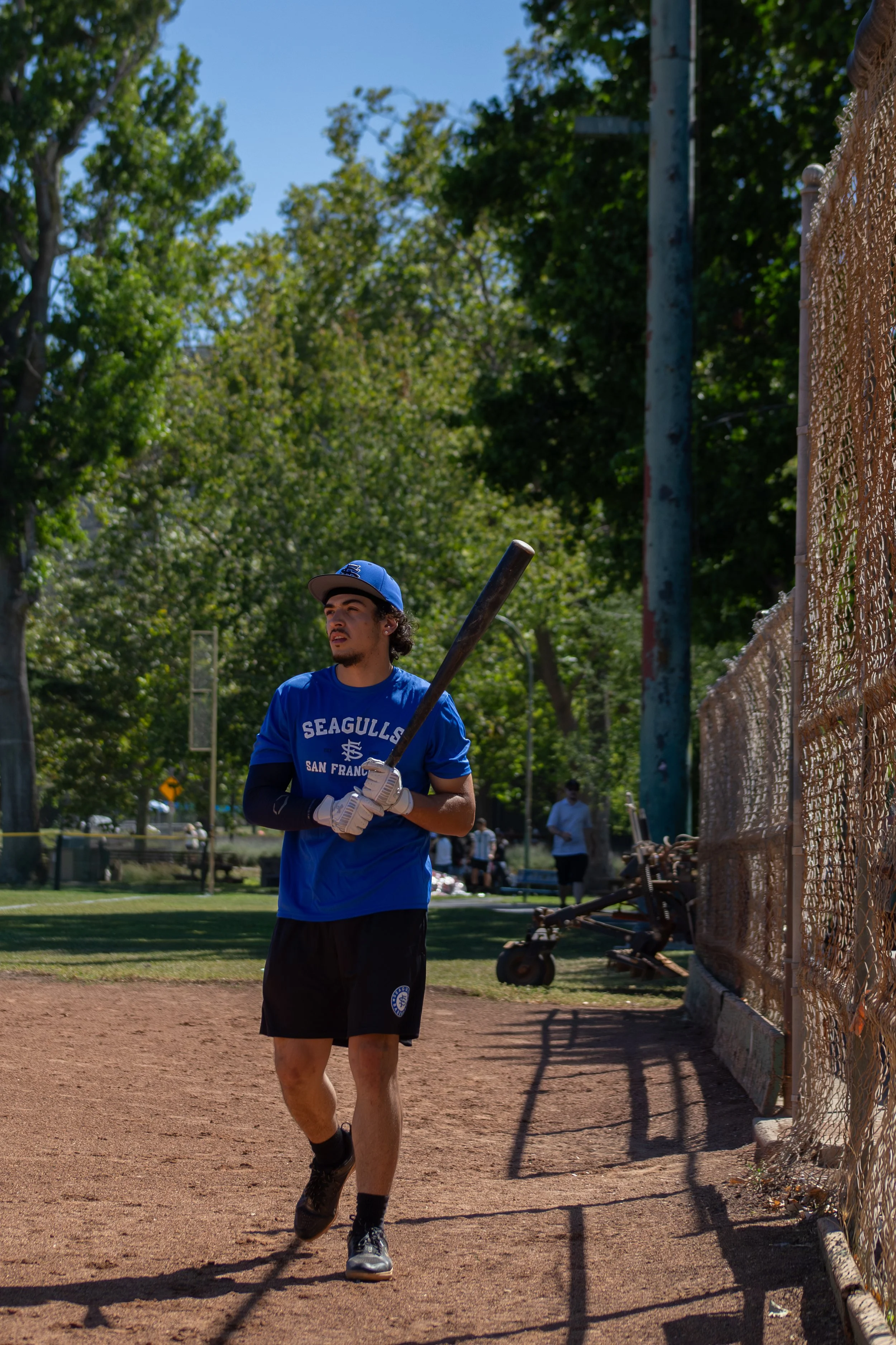 A young man wearing a blue baseball jersey and hat stands on a baseball field holding a bat, with a fence and trees in the background.