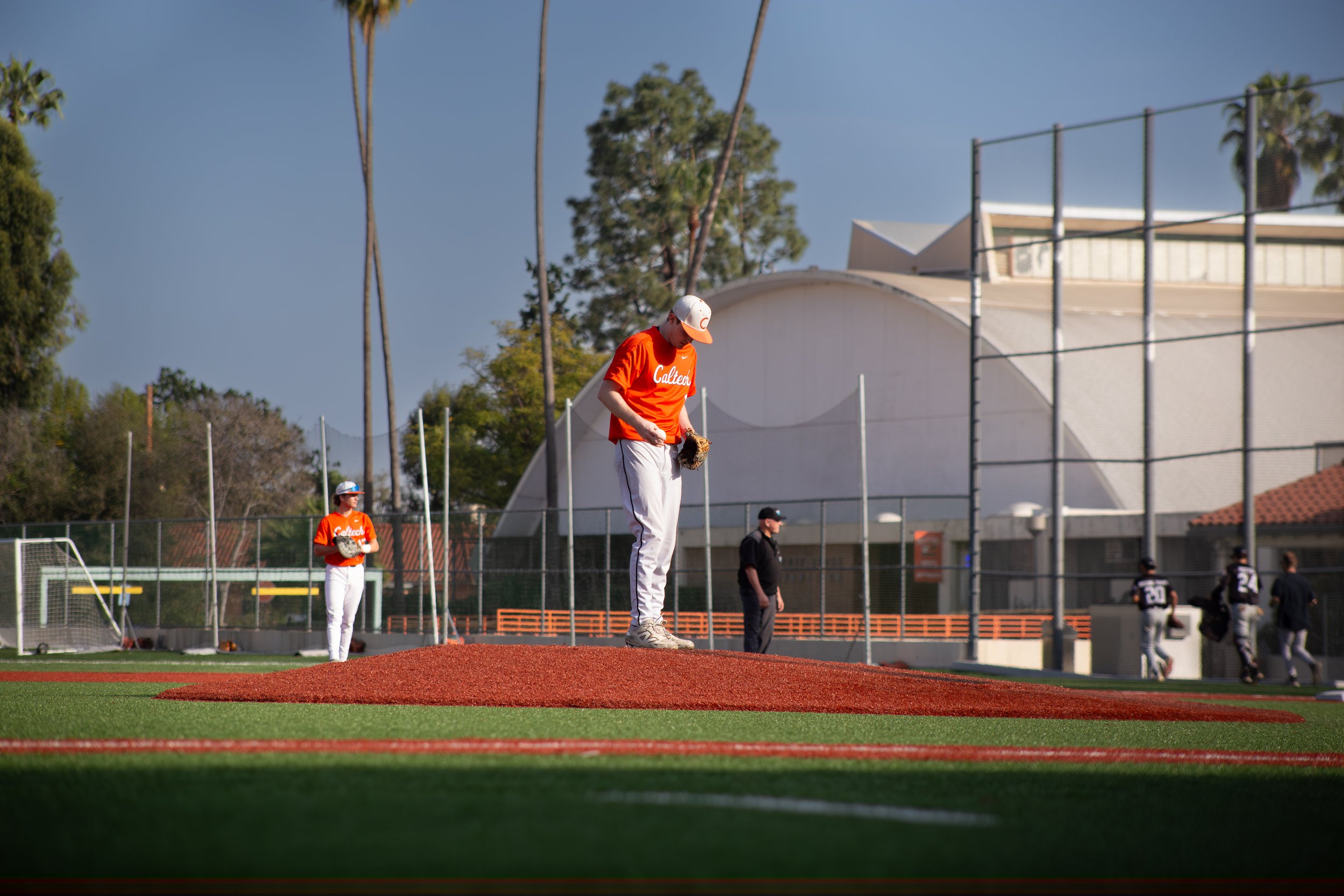 A group of baseball players on a field during practice, with one player standing on the pitcher's mound and others in the background, and a partly cloudy sky overhead.